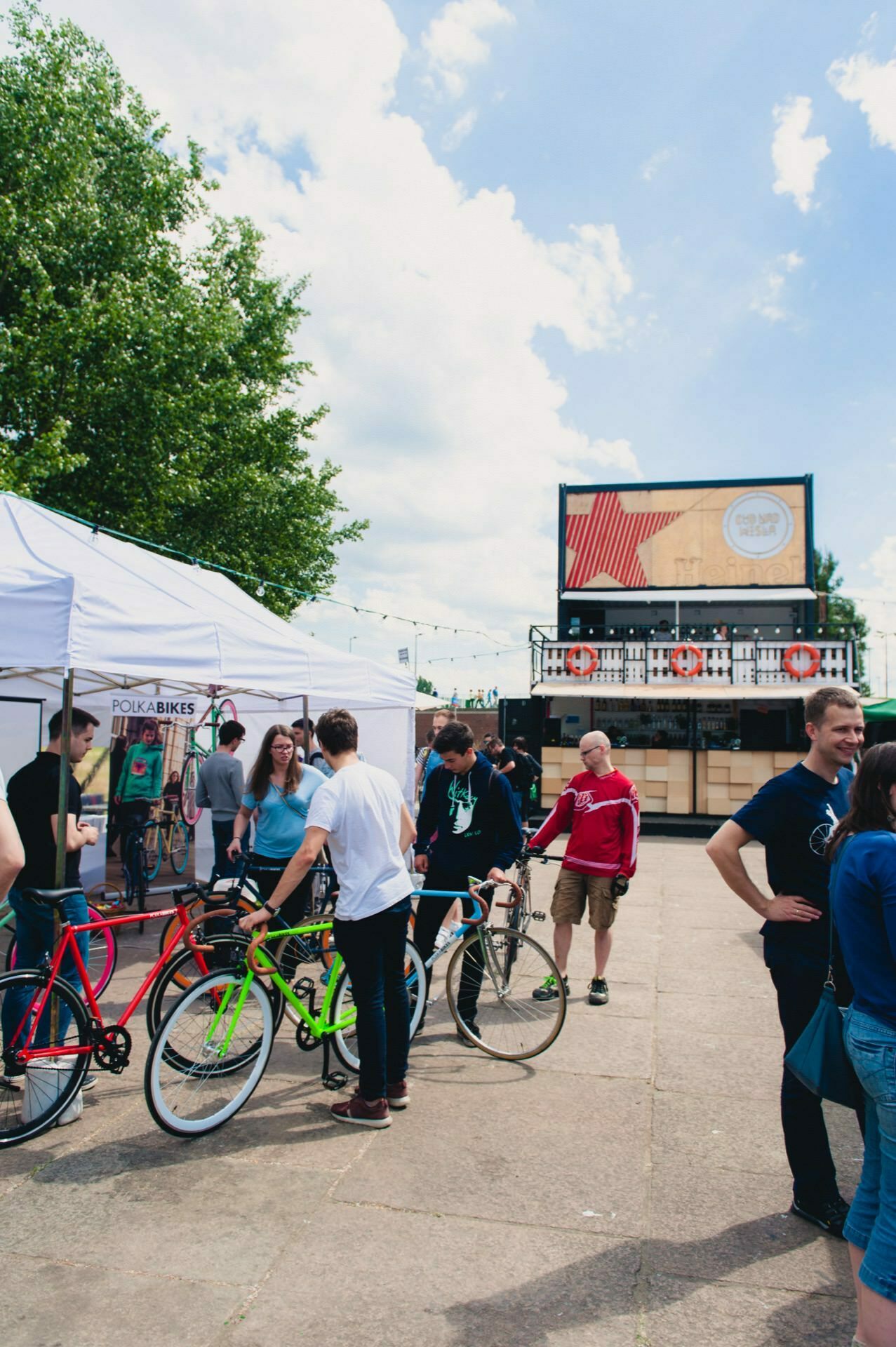 People gather around colorful bicycles under white tents at an outdoor event on a sunny day. Some people are talking or watching the bikes, others are passing by. A large sign or billboard and green trees are visible in the background, beautifully captured by the event photographer.  