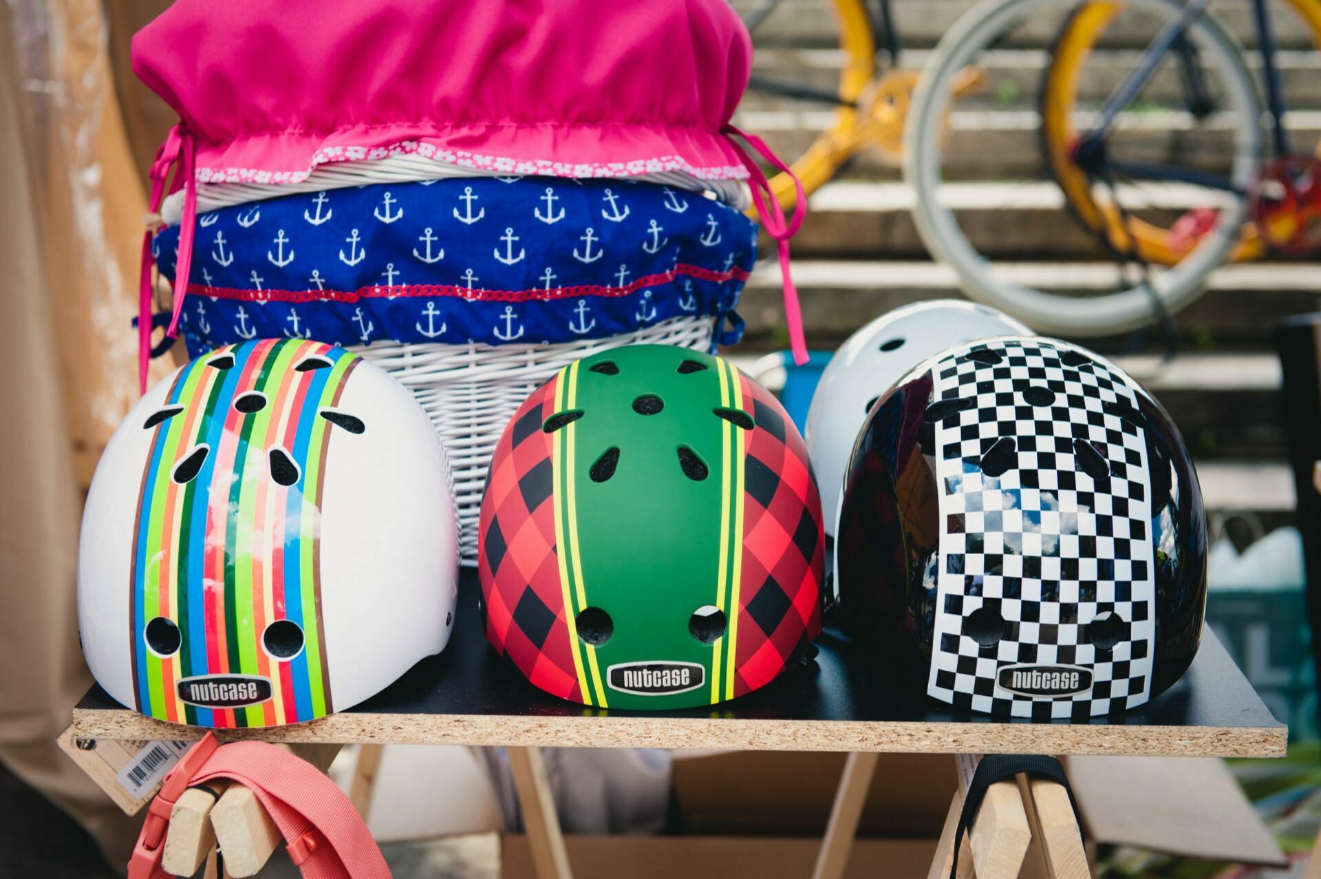 Four colorful bicycle helmets are lying on the table. Each helmet has a unique design: green stripes, red and green check, black and white check and multicolored stripes. Behind the helmets is a blue bag decorated with white anchor motifs, beautifully captured in this photo report from the fair.  