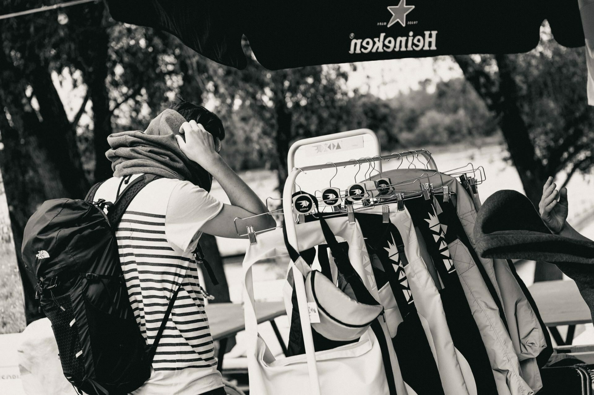 The black and white photo shows a person with a backpack browsing a stall filled with various hats and clothing hanging on a rack. They are mending a scarf around their neck in the shade of a Heineken-branded umbrella. This scene is part of an engaging photo essay of the fair, highlighting the atmosphere of the day.  