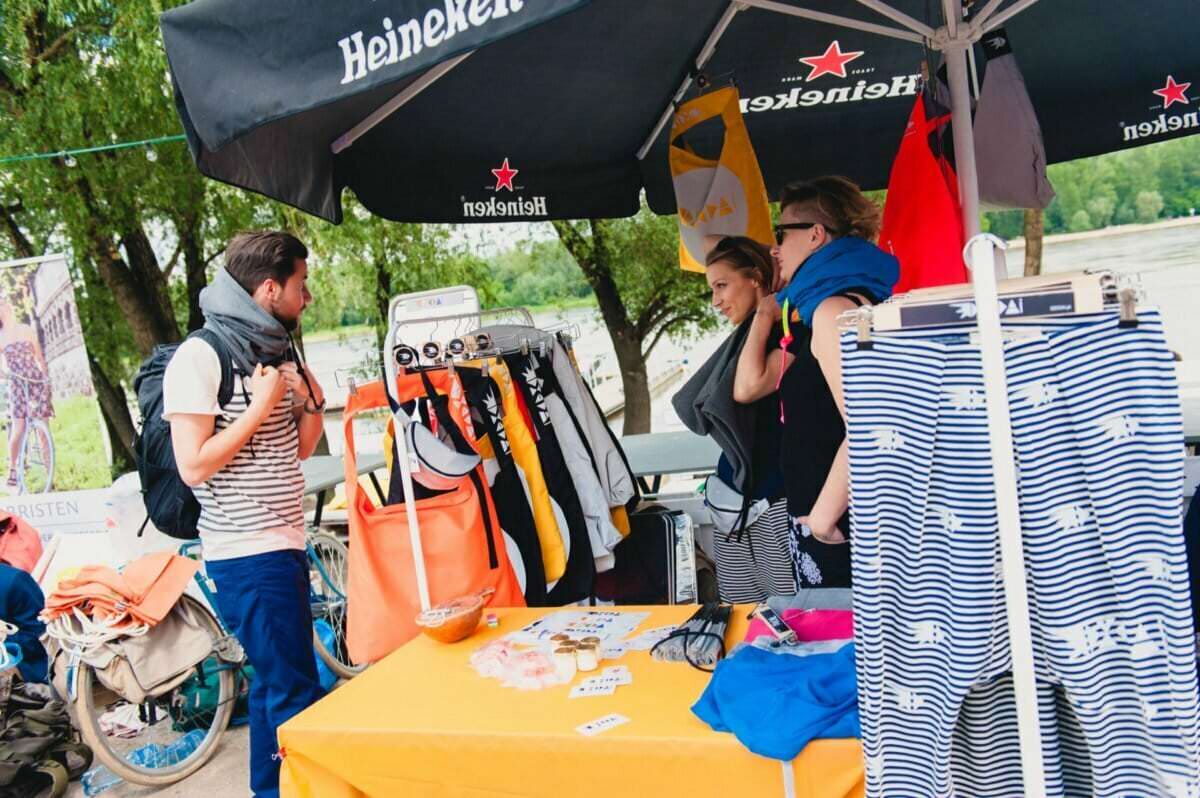 Trzy osoby przeglądają ubrania i akcesoria wystawione na żółtym stole i stojaku pod czarnym parasolem Heinekena na targu plenerowym. Tło stanowią drzewa, a atmosfera jest swobodna i zrelaksowana – idealna scena na fotorelację z targów.