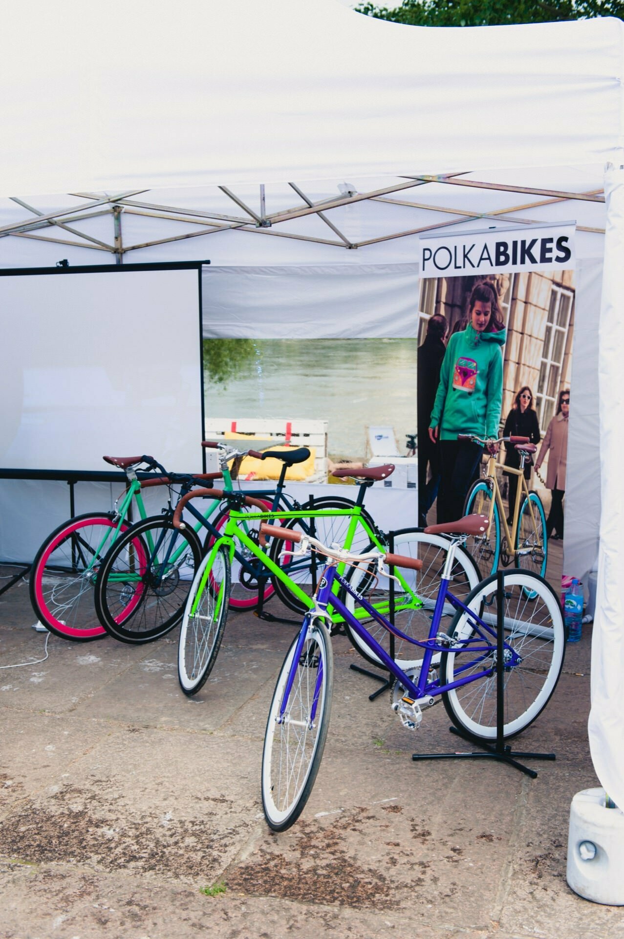 There are several colorful bicycles on an outdoor bike rack under a white canopy. A banner with the words "POLKA BIKES" and a blurred background showing people near the river is visible. White partitions, bicycle accessories and an event photographer capturing the moment complete the look, perfect for a photo essay of the fair.  