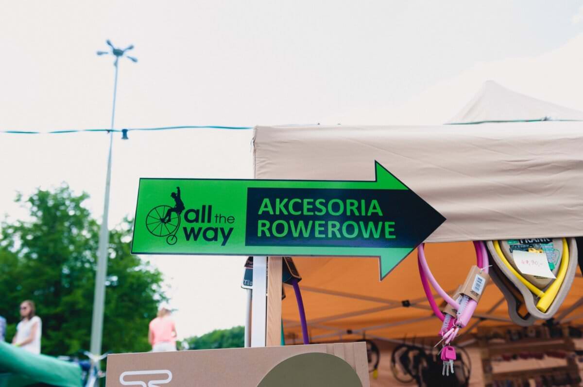 Zielony znak kierunkowy z czarną strzałką wskazuje w prawo i zawiera napisy „All the Way” oraz „Akcesoria Rowerowe”. Znak jest zamontowany na beżowym namiocie na targu na świeżym powietrzu i prezentuje znajdujące się w środku różne akcesoria rowerowe. W tle fotorelacji z targów widać drzewa i latarnie.
