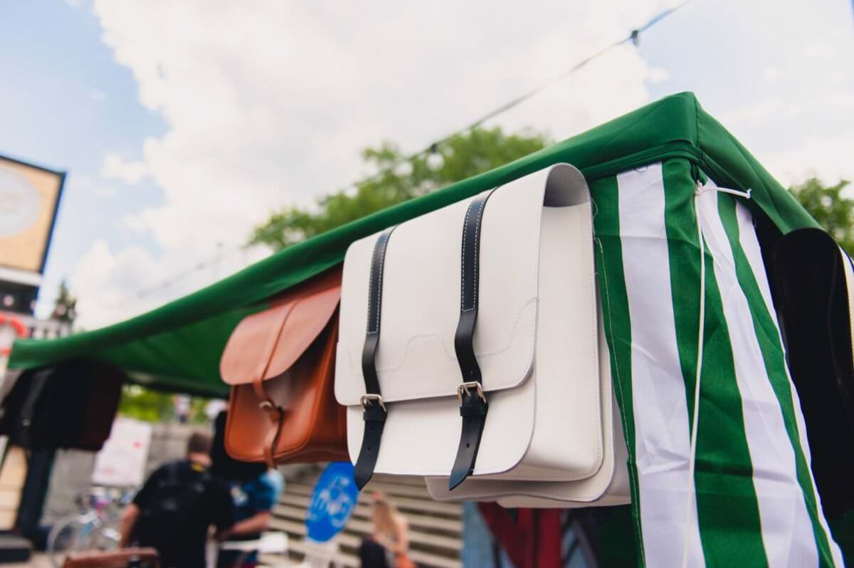 Zbliżenie na skórzane torby wiszące na wystawie na świeżym powietrzu na straganie z zielonym baldachimem, uchwycone przez fotografa eventowego. Centralny nacisk położony jest na białą torbę z czarnymi paskami. Inne torby w kolorze brązowym i brązowym są widoczne w tle, gdy ludzie przechadzają się po tętniącym życiem rynku.
