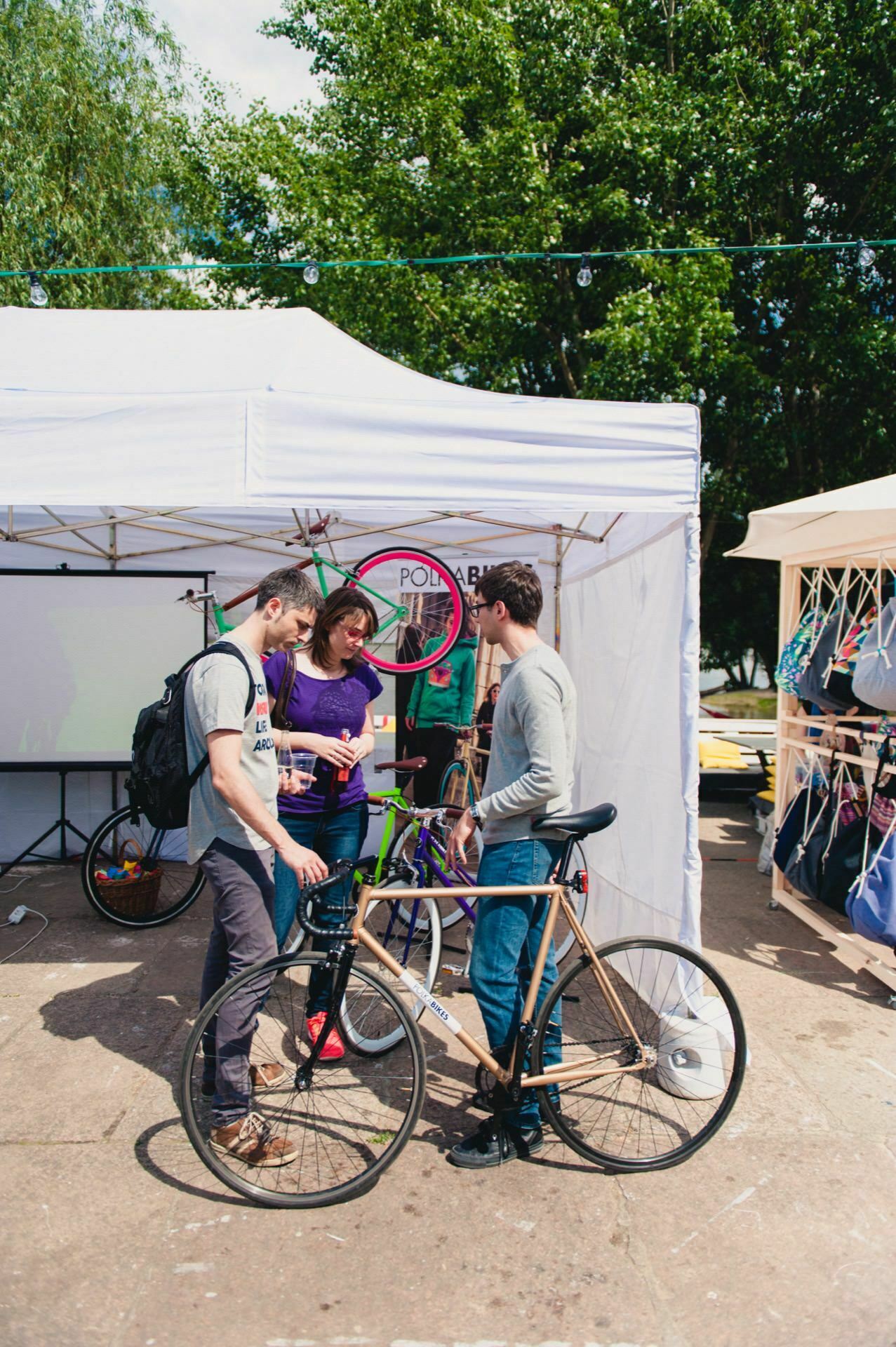 Three people stand near bicycles in front of a white tent during an outdoor event. Two men are facing a woman, who is probably holding a drink. A bicycle wheel hangs on the tent. The background captured as part of the photo report of the fair is greenery and other stalls.   