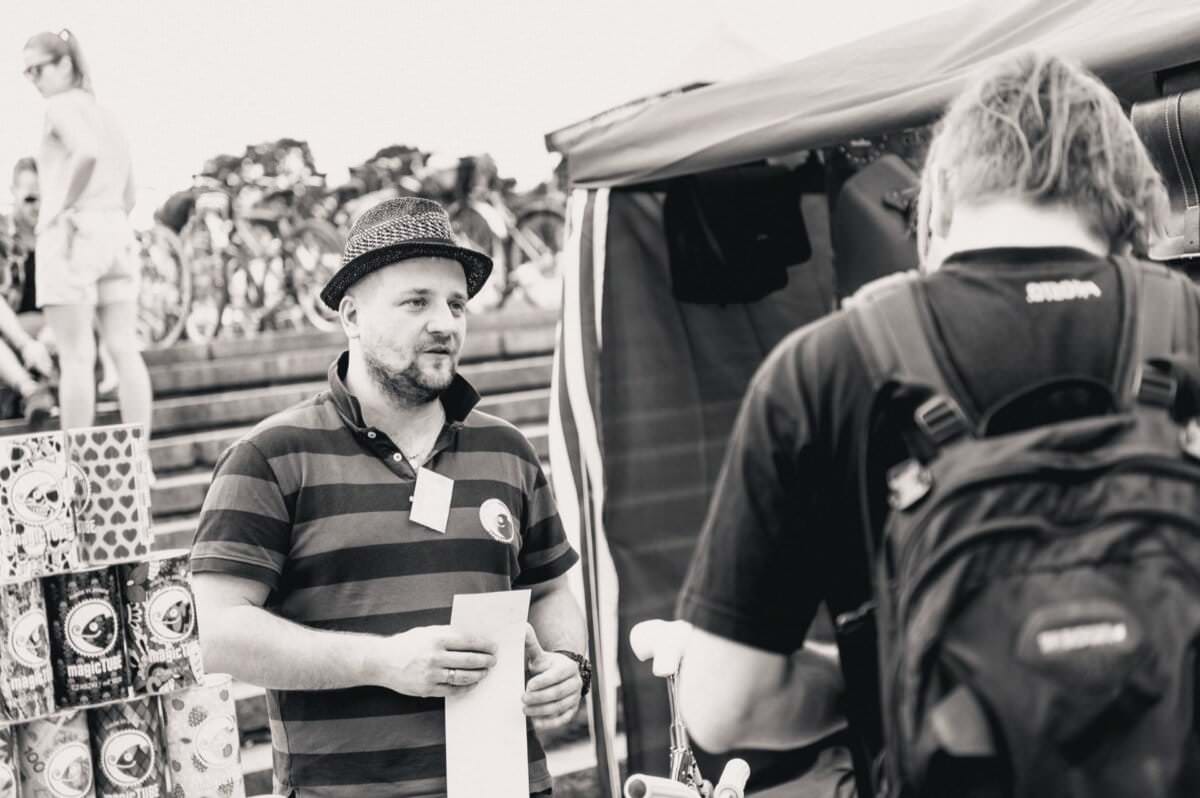 Mężczyzna ubrany w koszulkę polo w paski i kapelusz trzyma kartkę papieru, stojąc przed namiotem na targu na świeżym powietrzu. Naprzeciw niego stoi kolejna osoba z plecakiem. Po lewej stronie wyświetlane są różne produkty, a w tle rowery, pięknie uchwycone przez fotografa na wydarzeniu.