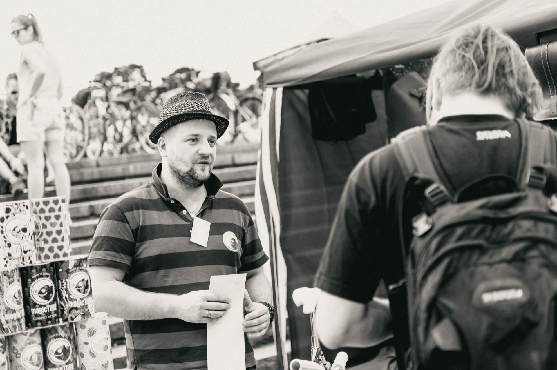 A man wearing a striped polo shirt and hat holds a piece of paper while standing in front of a tent at an outdoor market. Opposite him stands another person with a backpack. Various products are displayed on the left, and in the background are bicycles, beautifully captured by the photographer at the event.  