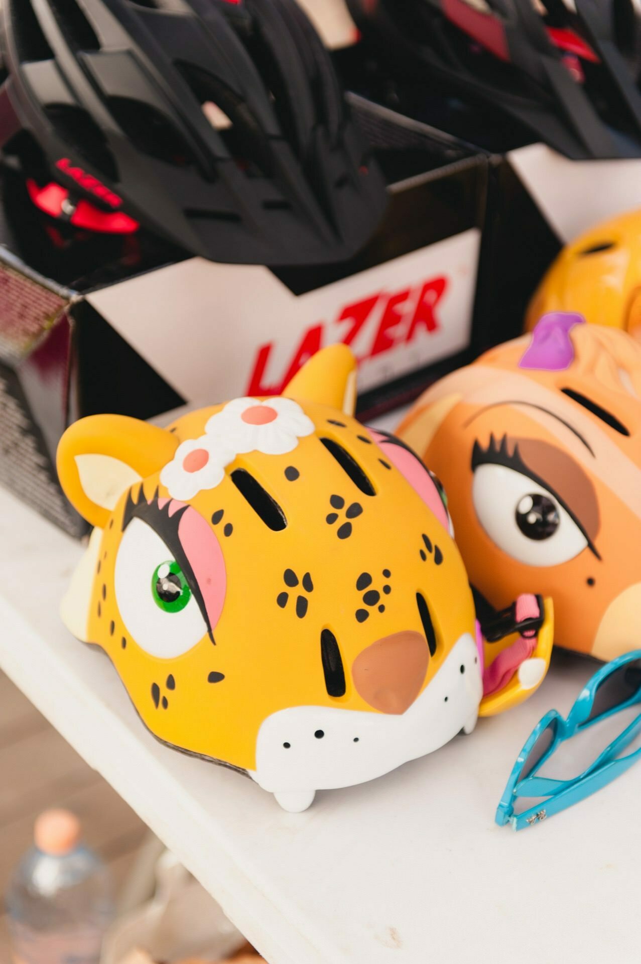 A table of colorful children's bicycle helmets was perfectly captured by a photographer from Warsaw. The closest helmet is bright yellow and has a cheetah face design, with green eyes and black spots. Next to it is another helmet with a tiger face design. Nearby are blue sunglasses.   