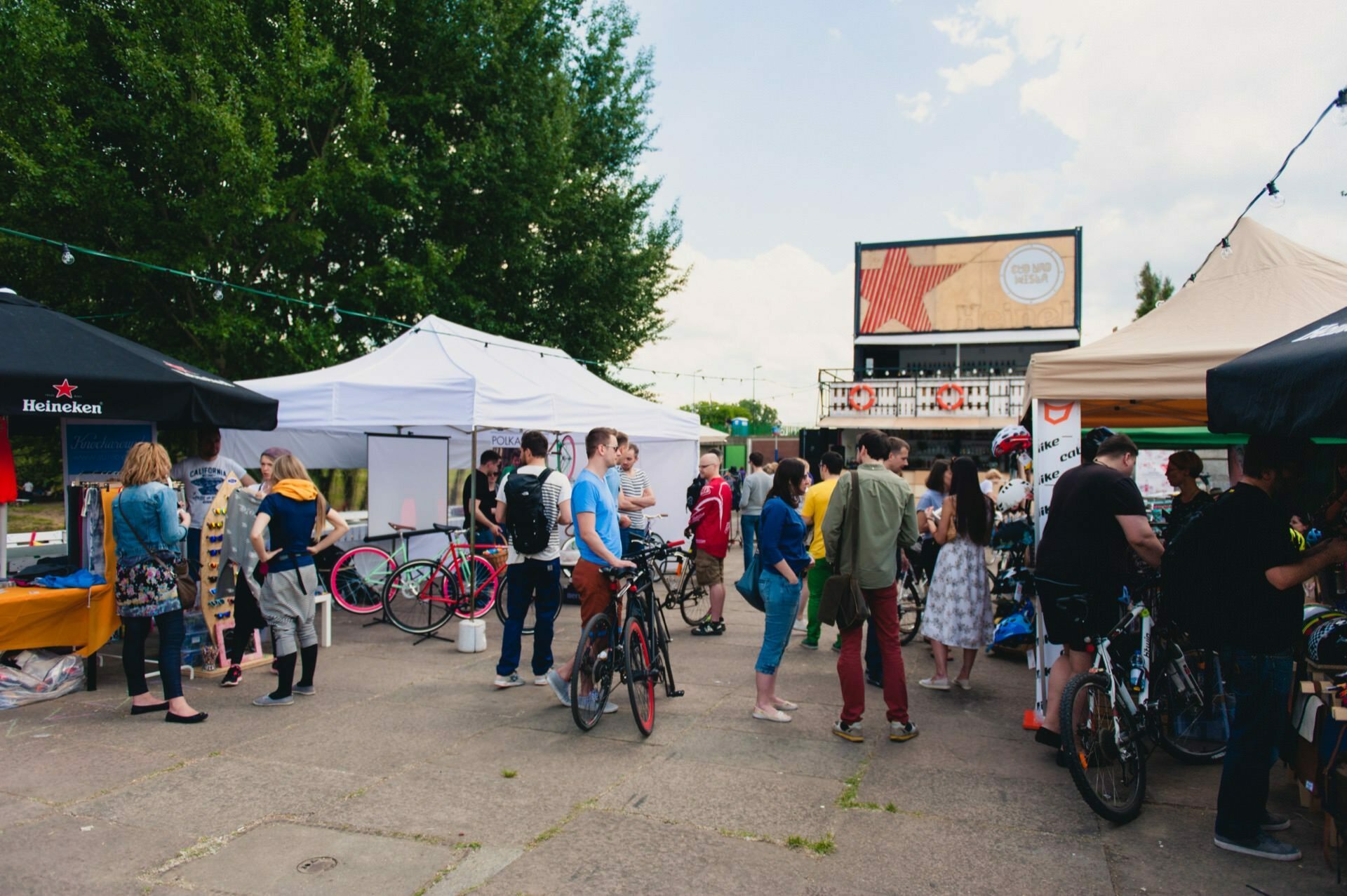 A bustling outdoor market with various stalls and tents where people browse items, ride bicycles and socialize. The scene includes a mix of trees and clear skies, all captured by a photographer from Warsaw. A large signboard is visible in the background.  