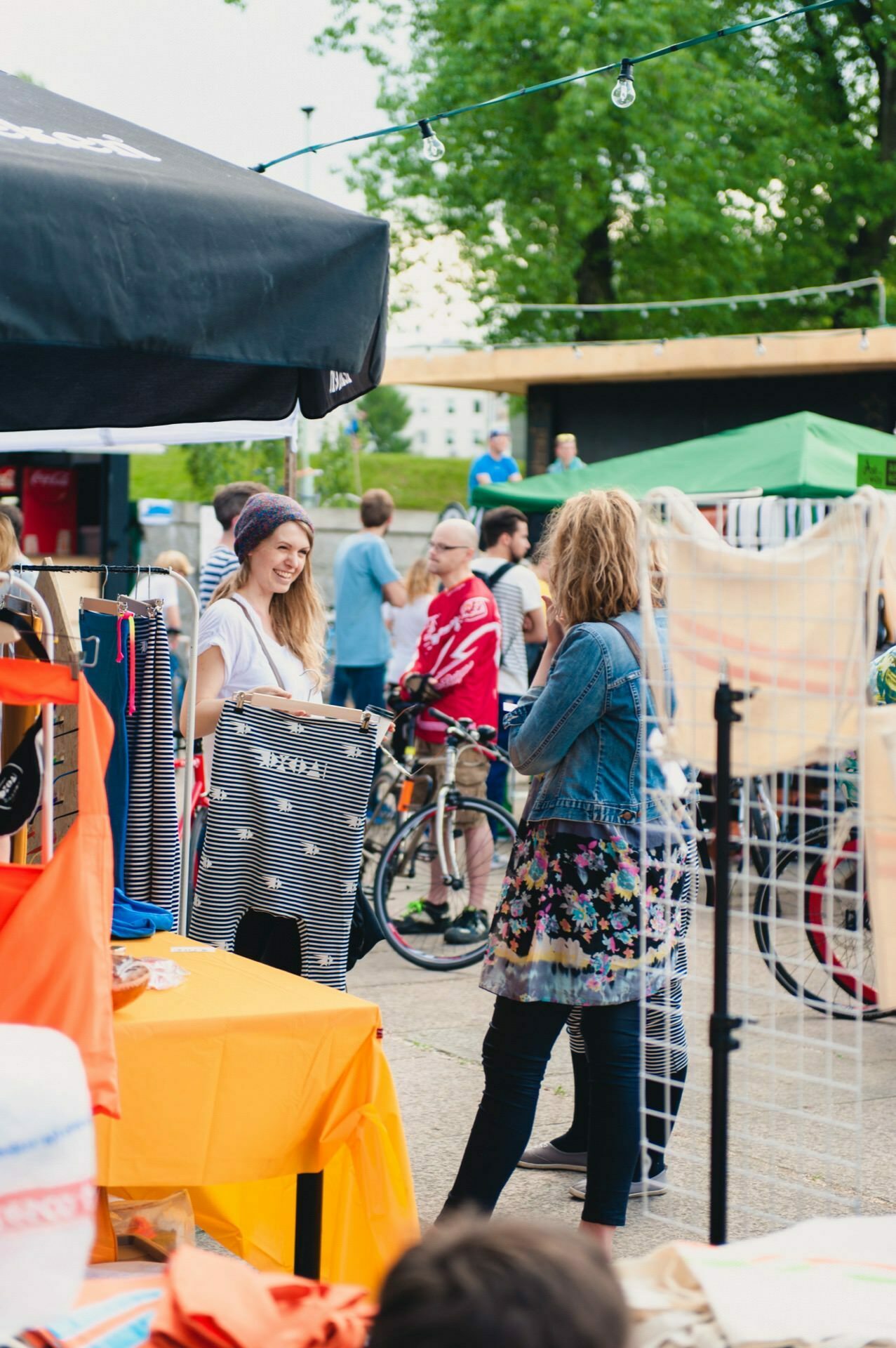 On a sunny day, an outdoor market is bustling with people browsing the stalls under tents. A woman wearing a cap and white shirt smiles and shows off her clothes to another woman in a denim jacket and floral dress, while a photographer at the event captures the bustling scene. Bicycles and trees can be seen in the background.  