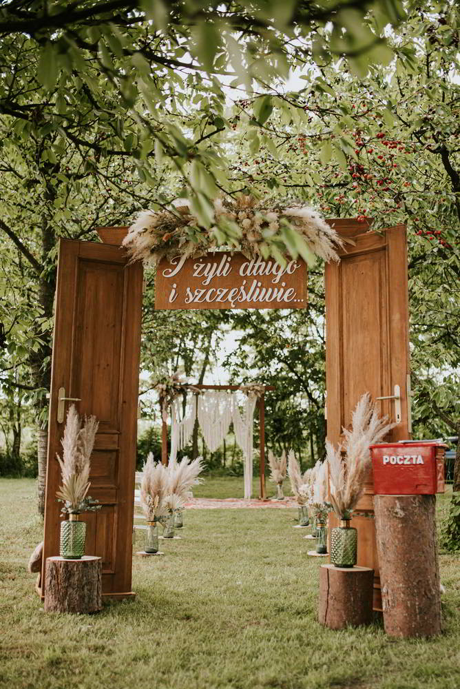 A rustic outdoor wedding arrangement with two wooden doors forming the entrance. Above, the inscription proclaims: "And they lived happily ever after," which in Polish means "And they lived happily ever after." A path planted with pampas grass leads to an altar under a tree canopy, beautifully captured by photographer in Warsaw Marcin Krokowski.  