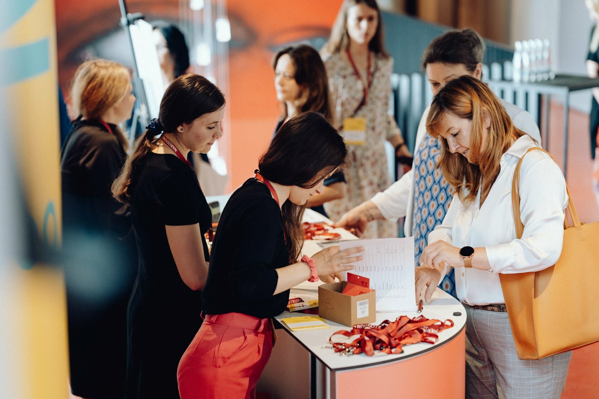 A group of women are standing around a table in what looks like a registration or check-in area for the event. Some are reviewing badges and lanyards, others are reviewing documents. They are in a modern area with bright lighting, perfect for any event photographer warszawa or event photographer.  