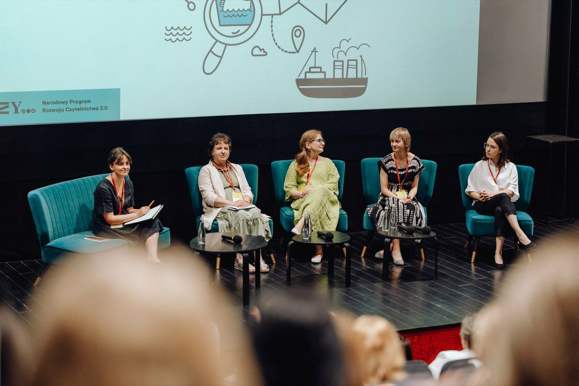 Five women sit on stage during panel discussions, seated on turquoise chairs, and a small table in front. Behind them is a large screen on which overview icons are displayed. The audience is visible in the foreground, and the entire scene has been beautifully captured by the photographer at the event, ensuring that every moment is documented.  