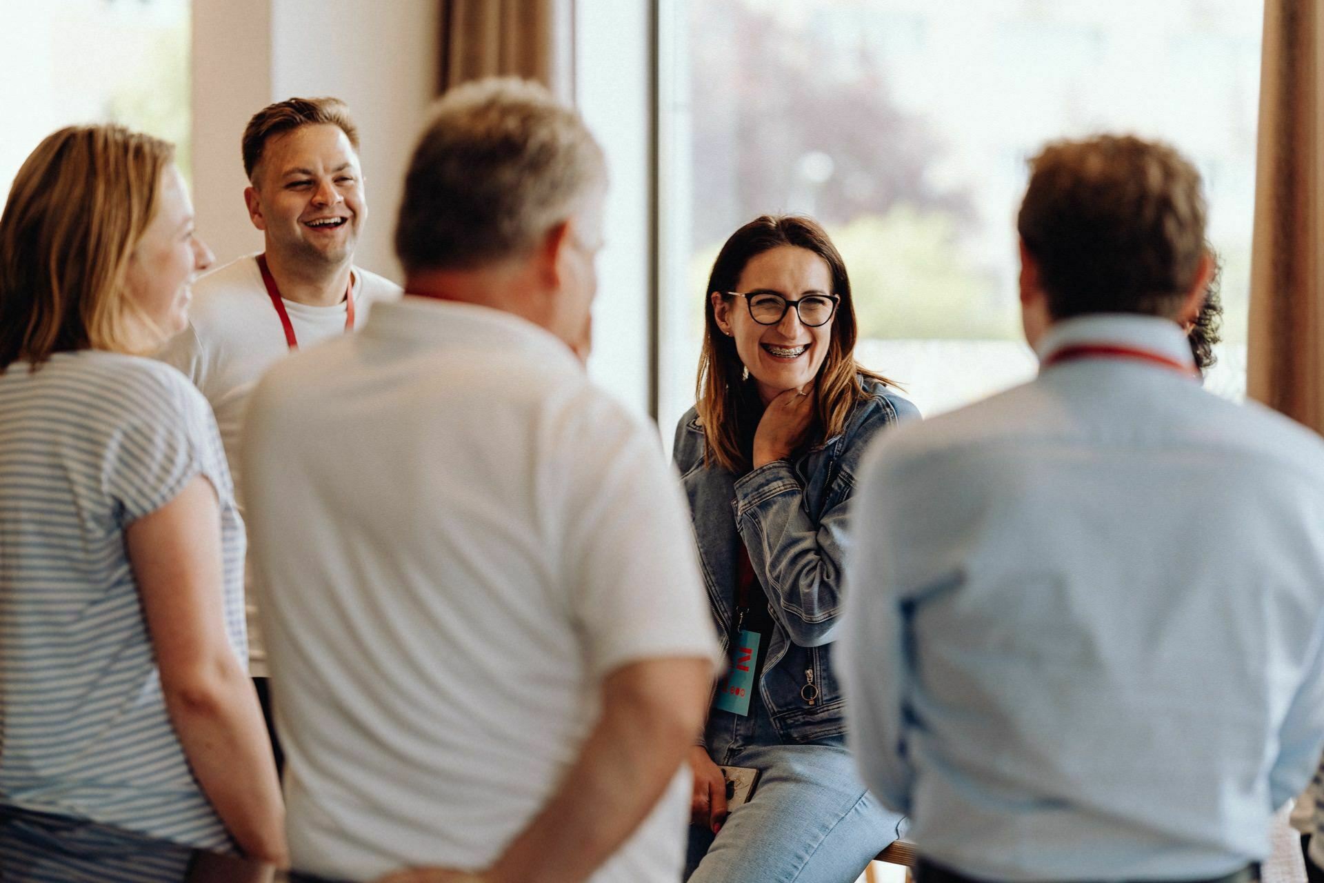 A group of five people, three men and two women, are having a lively conversation. One of the women is smiling broadly and resting her hand on her chin. It looks like everyone is enjoying the discussion in the bright room - which was perfectly captured by the photographer for the event.  