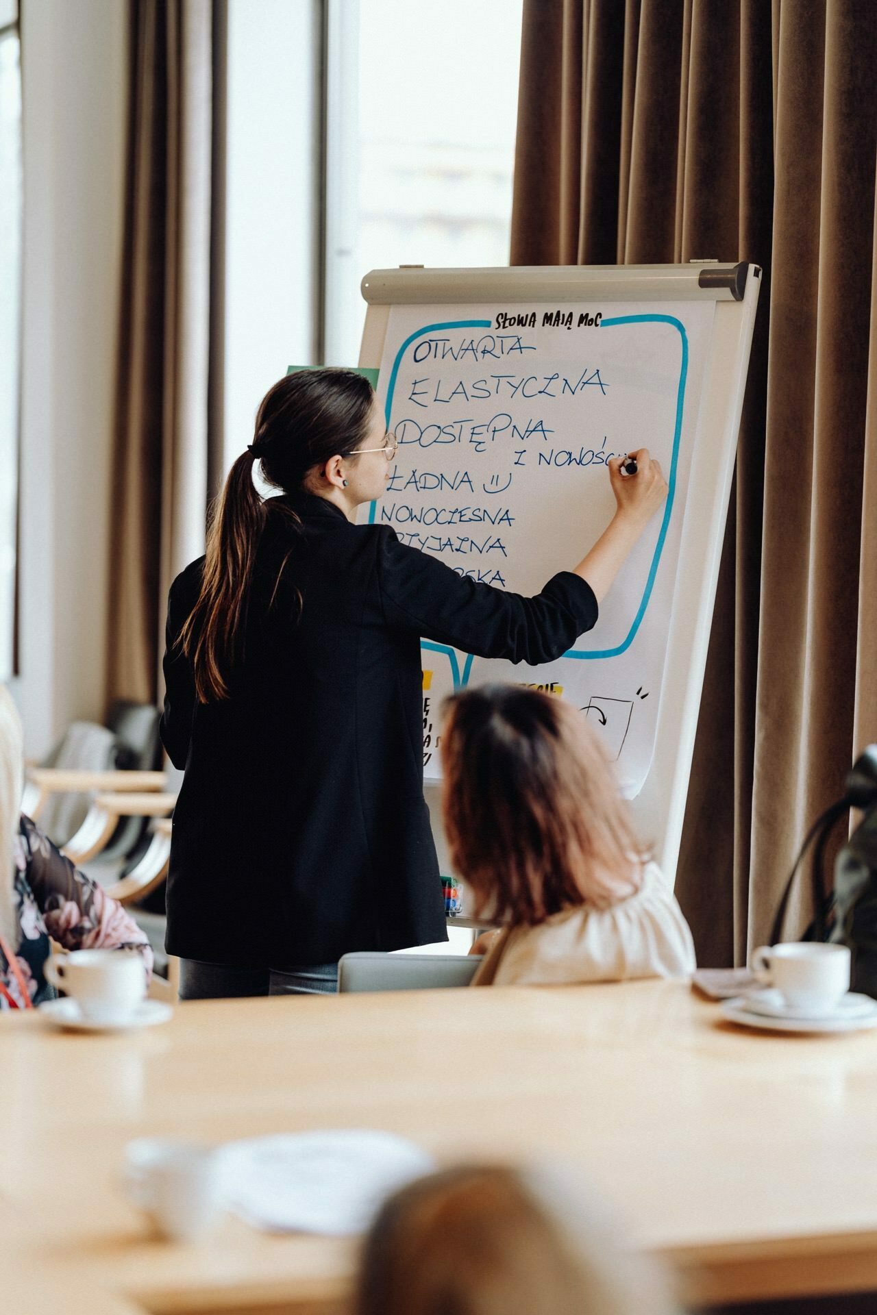 A woman in business attire writes during a meeting on a flipchart. The flipchart contains handwritten text in a foreign language. Several people sit around a large table and listen to her presentation. A photographer from Warsaw captures the moment in a room with large windows and beige curtains.   