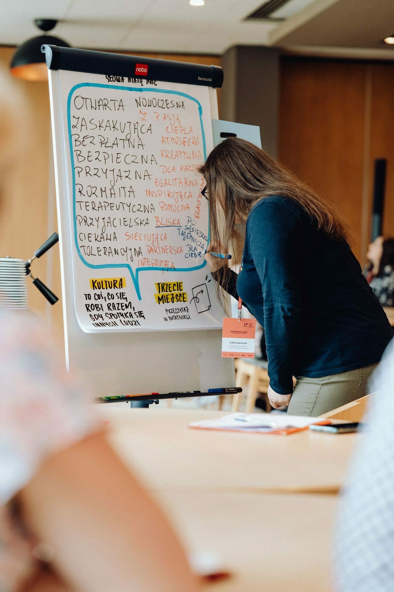 A person writing on a whiteboard filled with colorful words and phrases in Polish during a workshop or seminar. A person dressed in a dark blue top and sporting long brown hair was beautifully captured in a photo report of the event. The room has a wooden interior, and other participants are present in the background.  