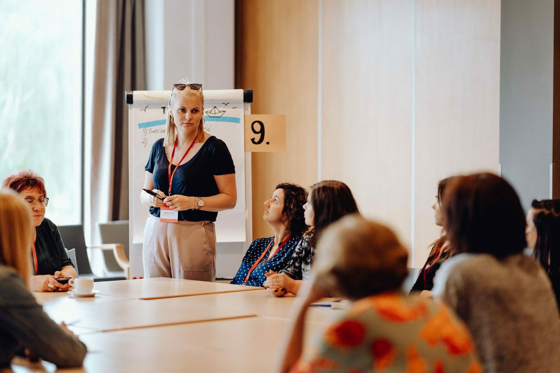 A woman stands next to a flip chart in a conference room and speaks to a group seated around a table. The participants, wearing badges, listen attentively, joining in the discussion. The setting looks professional, and the room is illuminated by natural light - a perfect scenario for a photo event.  