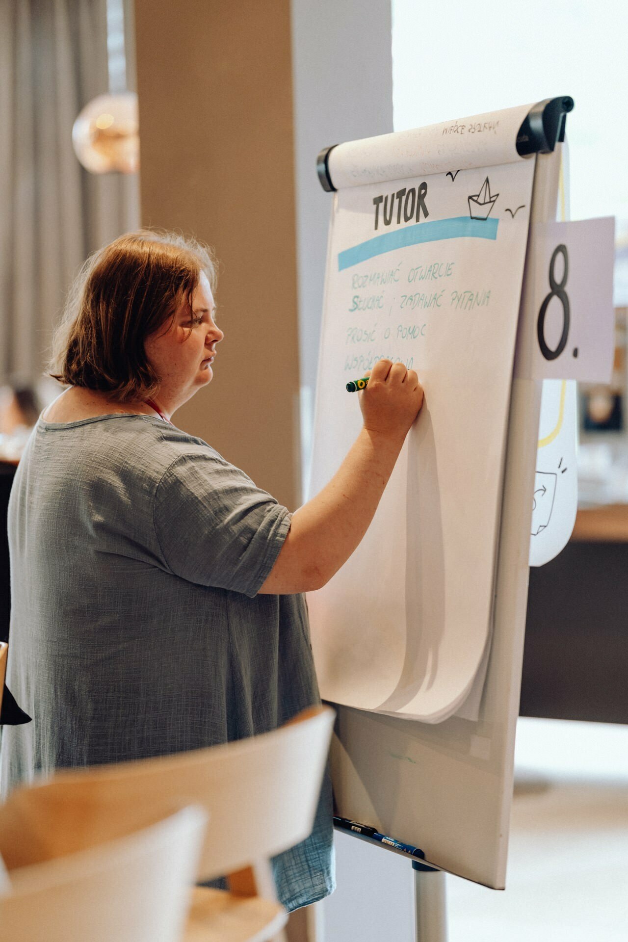 A person with shoulder-length hair stands next to an easel with a large notepad and writes with a marker. The top of the easel reads "TUTOR," with a few partially visible words underneath. The person, presumably captured in *event photography warsaw*, is focused on writing and gives the impression of being in a classroom or meeting.  