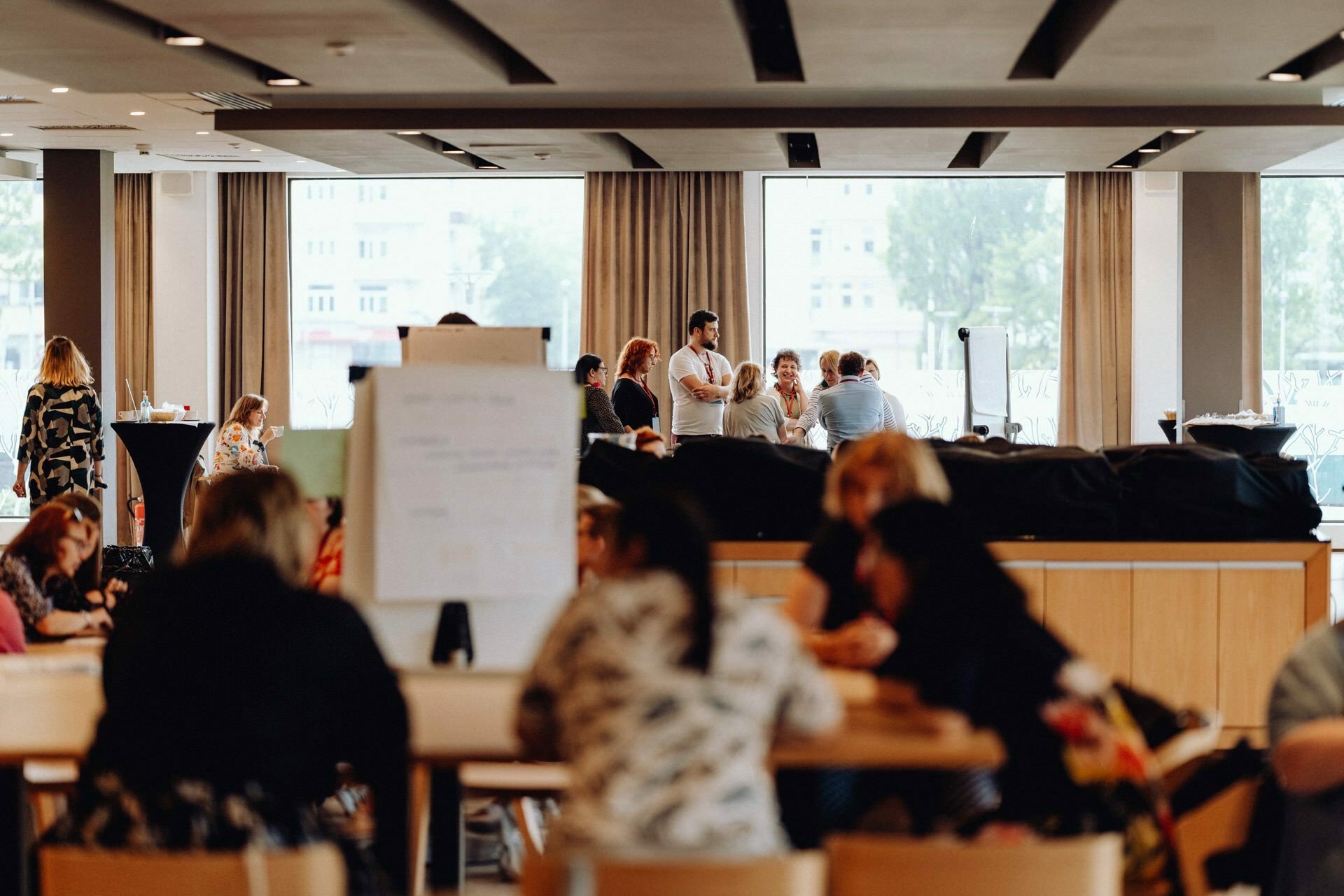 A group of people are discussing and collaborating in a spacious, modern room with large windows. Some sit at tables, others stand in groups. The atmosphere seems to be a mix of a casual meeting and a brainstorming session, perfect for an event photographer in Warsaw to capture the moments.  