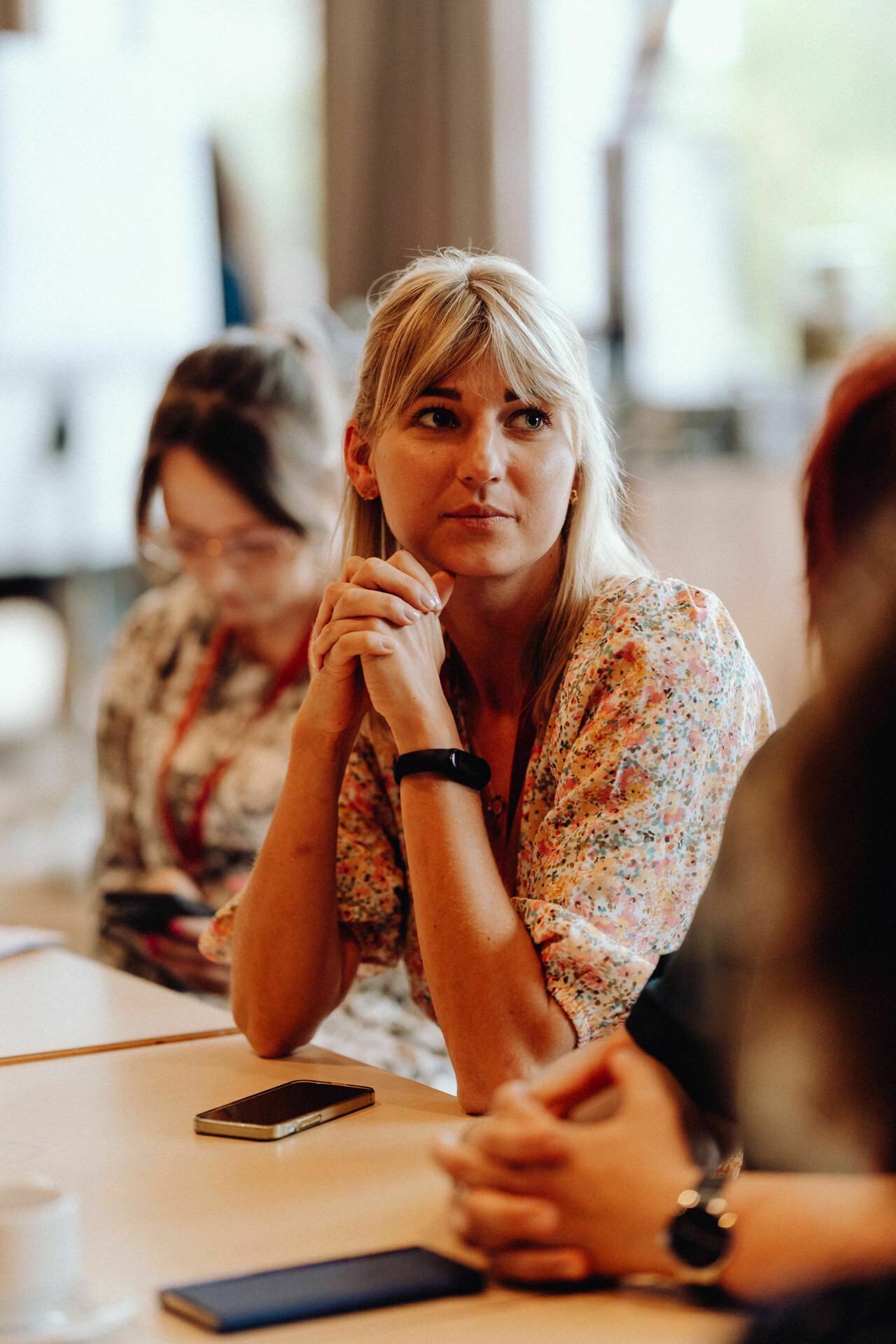 A woman with blonde hair, wearing a patterned blouse, sits at a table with her hands folded and stares intently. Next to her sits another woman and looks at her phone. The scenery is reminiscent of an ordinary meeting or discussion in a room, perfectly captured for the event photo report by a skilled event photographer.  