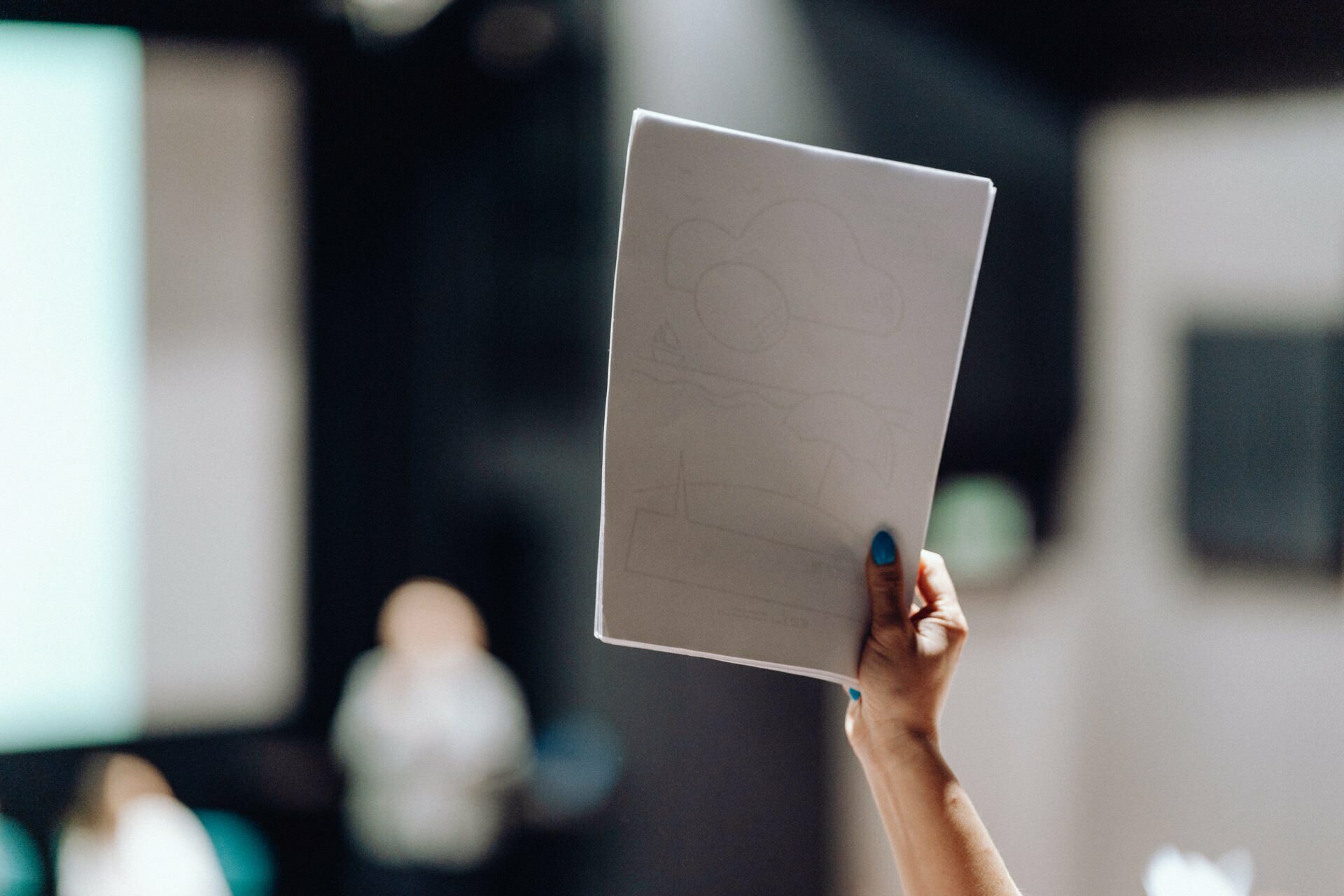 A person with blue nail polish is holding a piece of paper with a simple drawing of clouds and a landscape in a blurry room. The background is out of focus, suggesting that the person may be in an audience or classroom, which may have been captured by an event photographer Warsaw. 