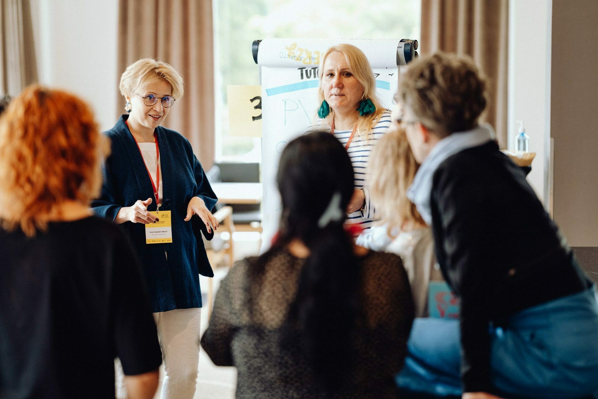 A group of people are having a discussion in a bright room with large windows. Two women stand at a whiteboard with notes and lead the conversation. Other participants, sitting or standing, listen attentively. Documents and markers are visible, capturing the essence of the event's photo-report.   