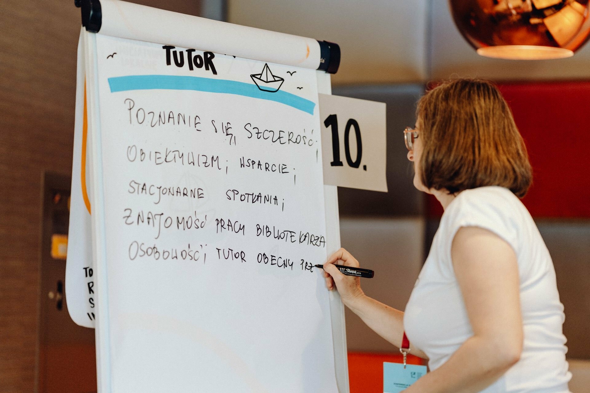 A person wearing glasses and a white shirt writes on a flipchart with the word "Teacher" written on it and decorated with a paper drawing of a boat. The chart contains handwritten notes in a language other than English. On the right side of the chart, a "10" sign is visible, beautifully captured by a photographer from Warsaw.  