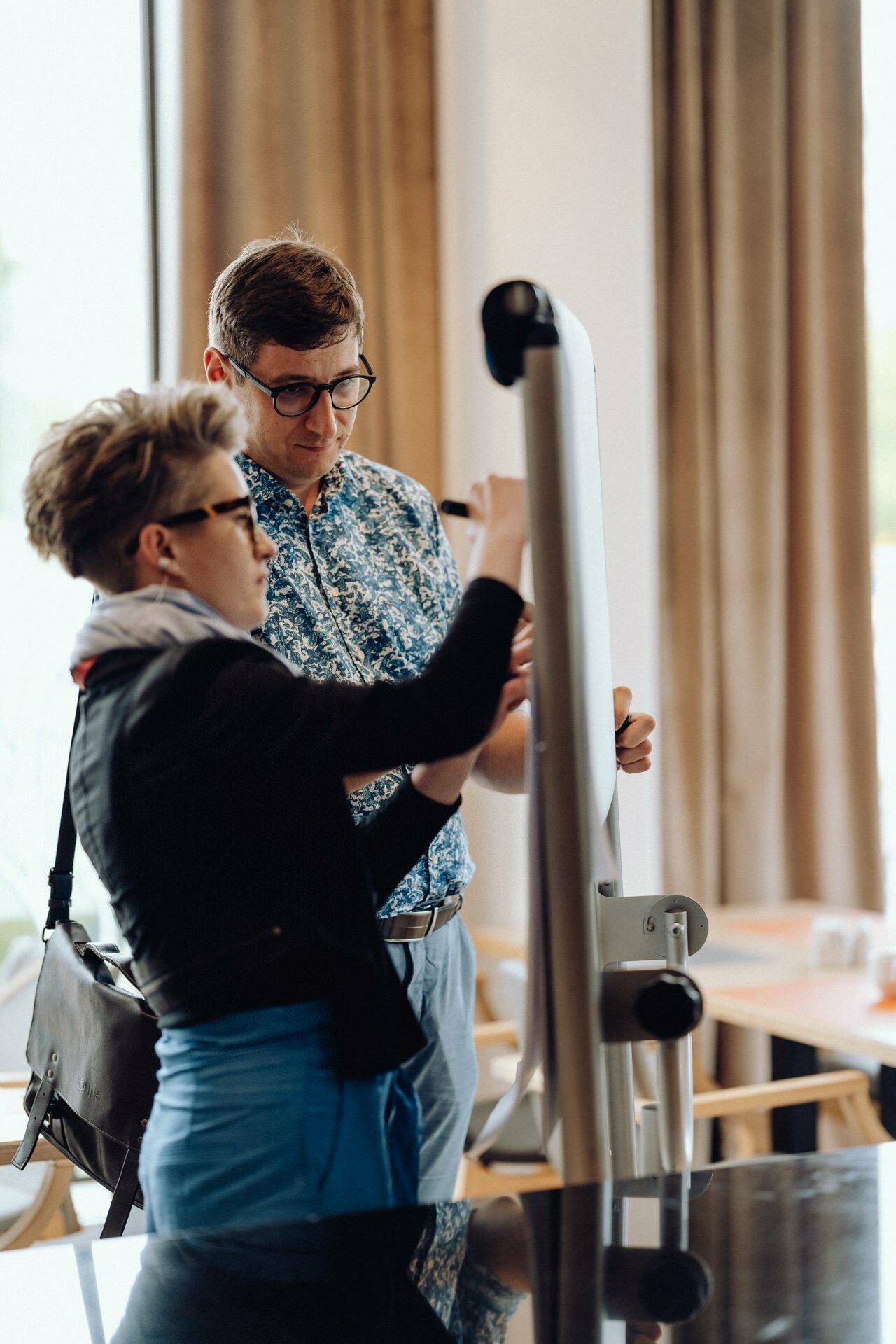 Two people, one wearing glasses and a floral shirt and the other in a black jacket and scarf, stand in front of a flipchart and write or point to something on it. A brightly lit room with large windows provides the setting for this photo report of the event. 