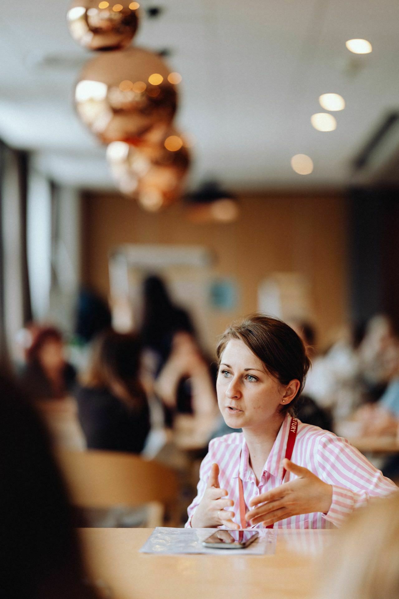 A person sits at a table and gestures with his hands as he speaks, in a busy room with large windows. The background is blurred, showing other people and round copper pendant lights hanging from the ceiling, perfectly captured by the event photographer Warsaw. 