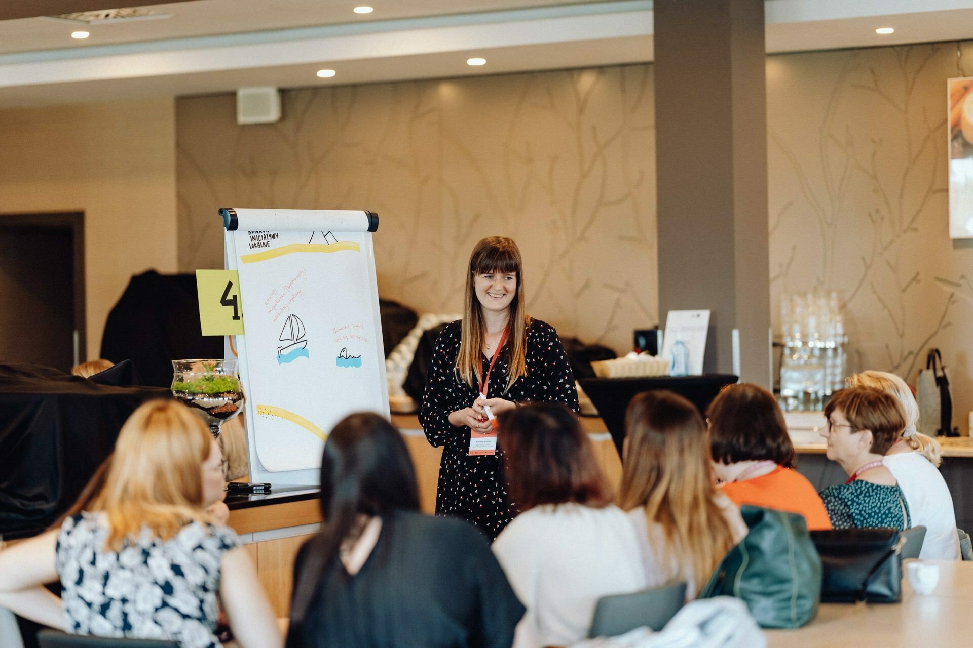 A woman stands and speaks in front of a group of seated people during a workshop. Behind her is a flipchart with colorful drawings. The participants are engaged and listening intently. The setting appears to be a casual, modern conference room, beautifully captured by a photographer for an event from Warsaw.   