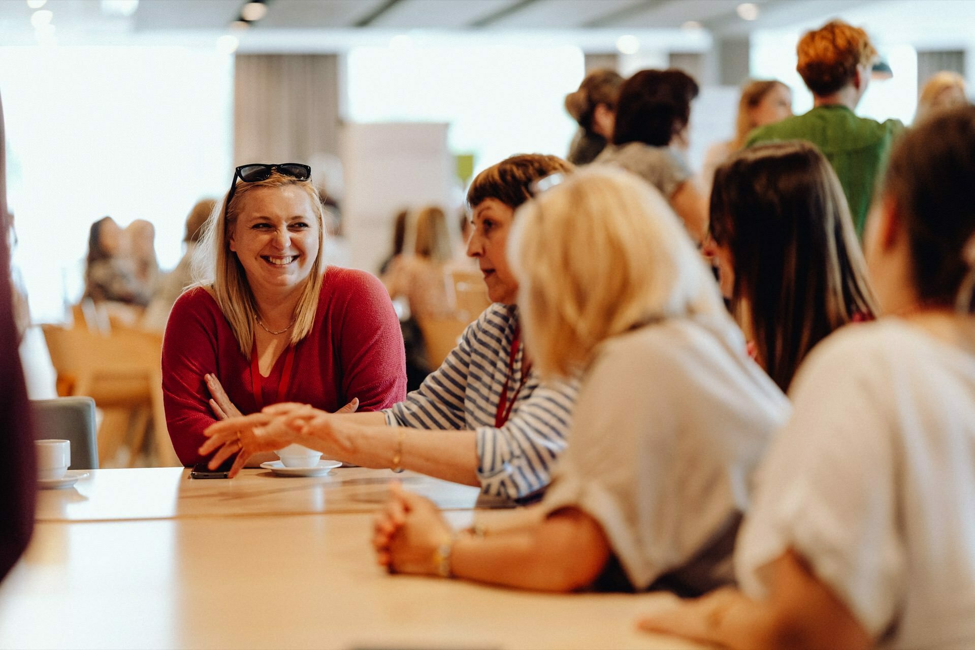 A group of people sitting around a table, immersed in conversation and smiling. The venue appears to be taking place in a closed room, probably a conference or social gathering. In the background, where a Warsaw event photographer might have captured a snapshot of the event, more people can be seen standing and talking.  