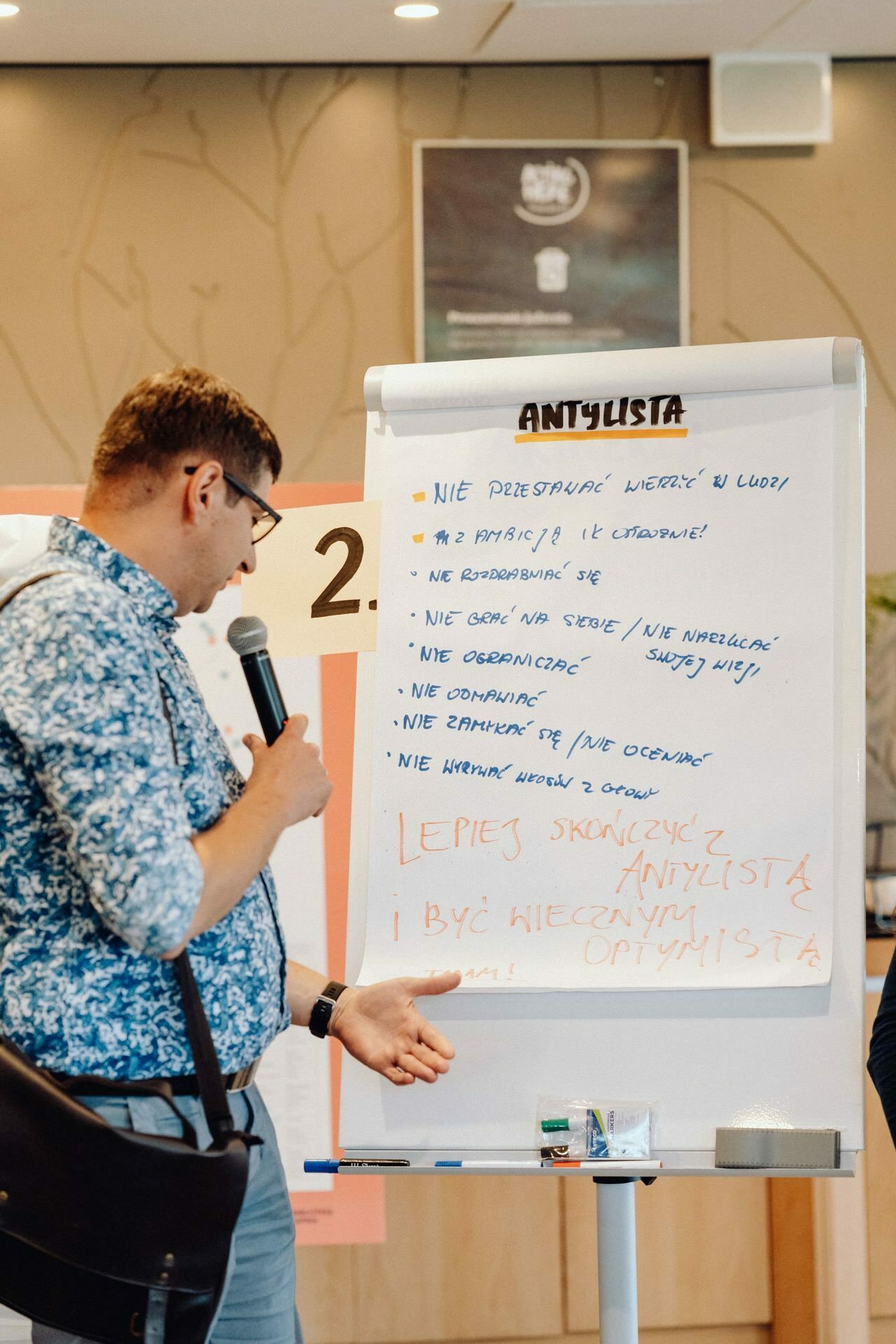 A man in a blue patterned shirt, holding a microphone, points to a flipchart with handwritten text. The heading reads "Anulista" and contains several numbered bullet points. The man is wearing glasses and has a bag slung over his shoulder. The blurry background captures the elegance of the event photo essay.   