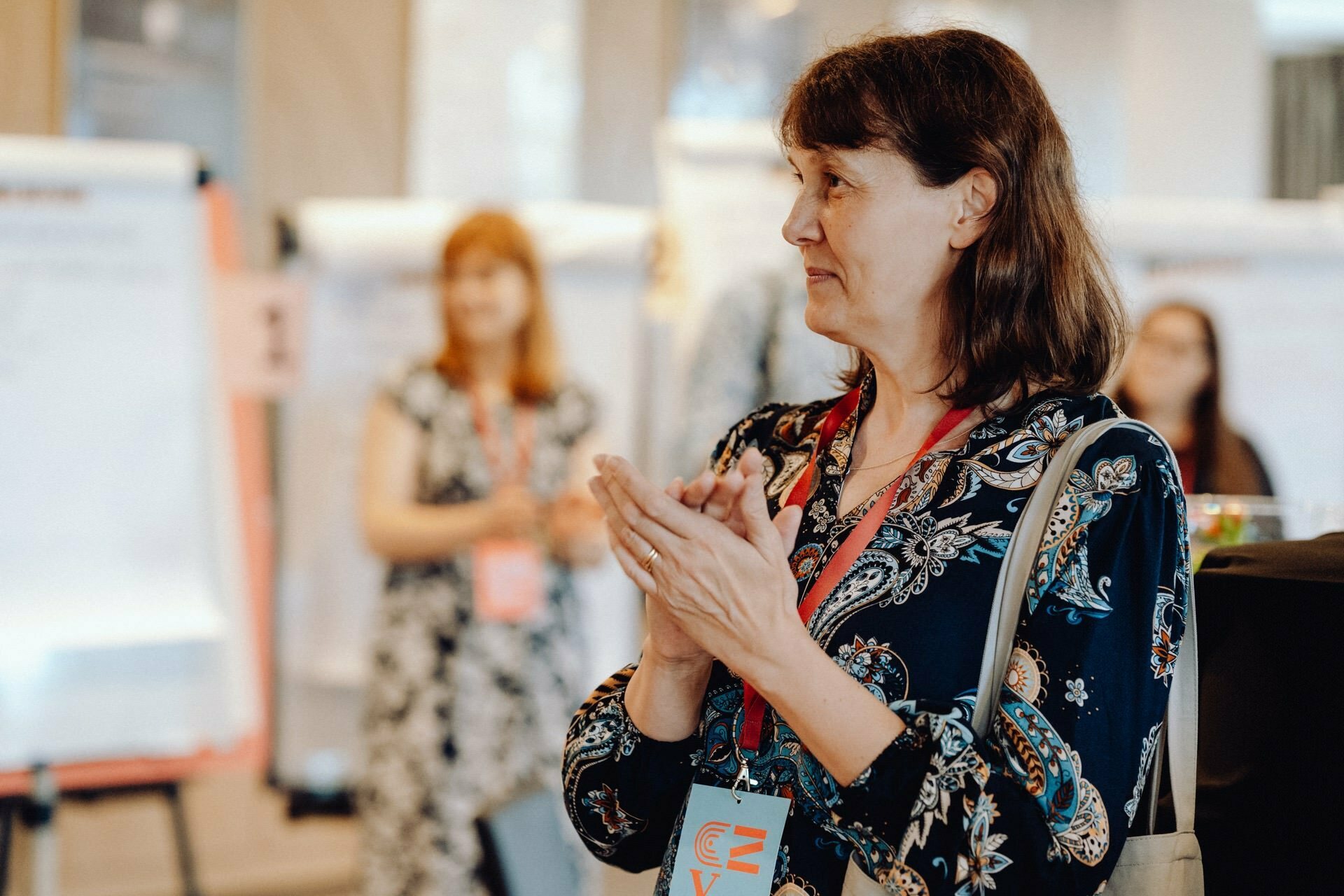 A woman with brown hair, wearing a dark blouse with flowers and a name badge, claps her hands in the room. In the background you can see people and large paper blocks on easels, suggesting a workshop or seminar. This scene captures the essence of the event's vibrant photo essay.  