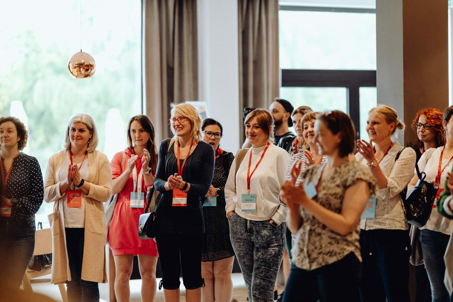 A group of people, mostly women, stand together in a room and clap, looking engaged and positive. Many wear name badges and lanyards. The scenery with large windows and natural light creates a friendly atmosphere - a perfect photo report of the event by a talented photographer from Warsaw.  