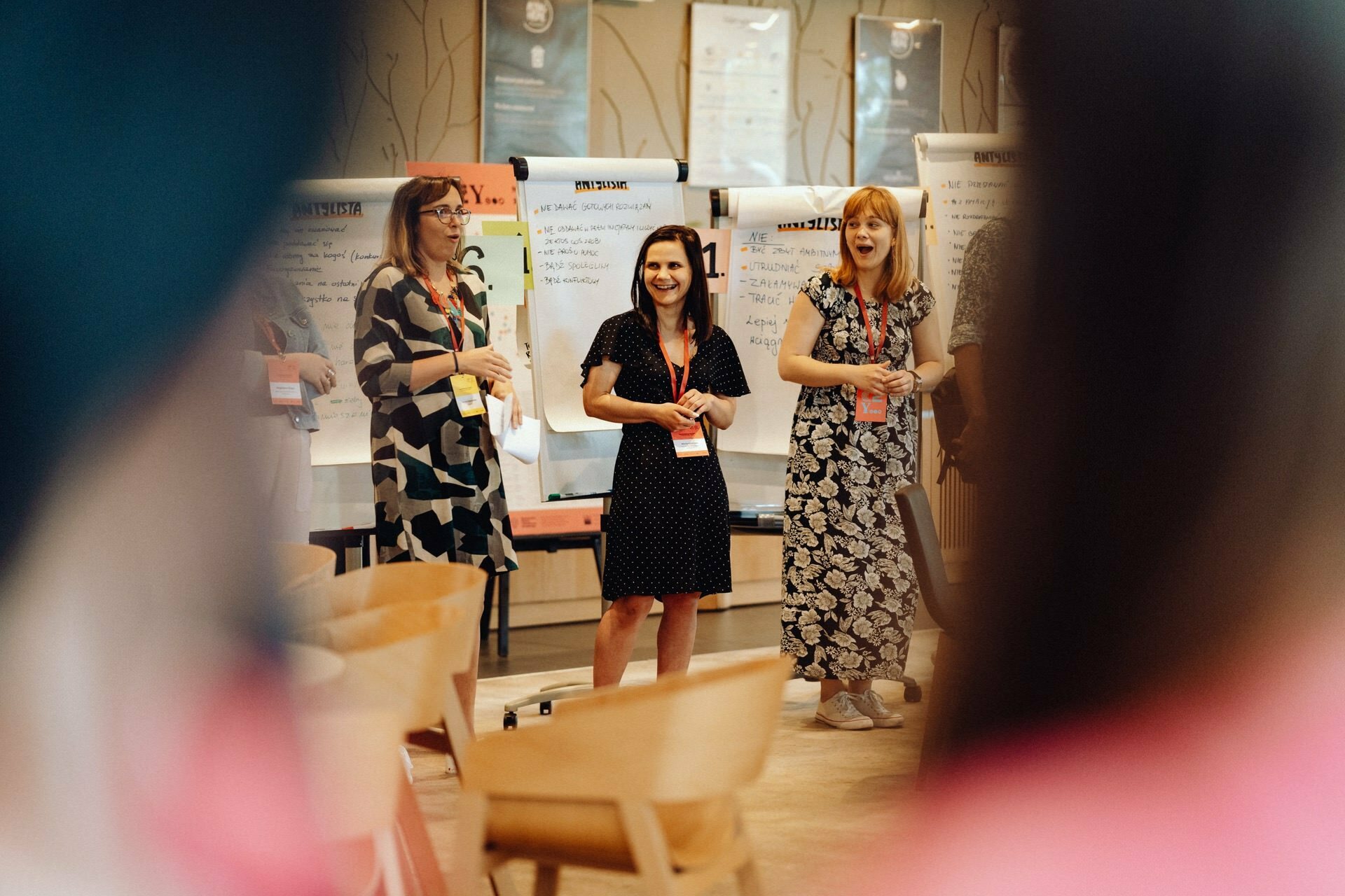 Three women stand and smile in front of flip charts in a brightly lit room, capturing a moment during a workshop or presentation. Chairs and additional people are blurred in the foreground, a perfect framing of the scene for a photo essay of the event. 