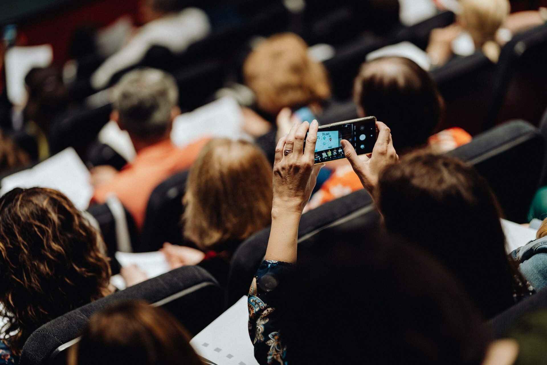 A person takes a picture with a smartphone in a crowded room, photographing people sitting in rows facing forward. Some people are holding documents suggesting that a lecture or presentation is in progress - the perfect event photo reference for any photography event. 