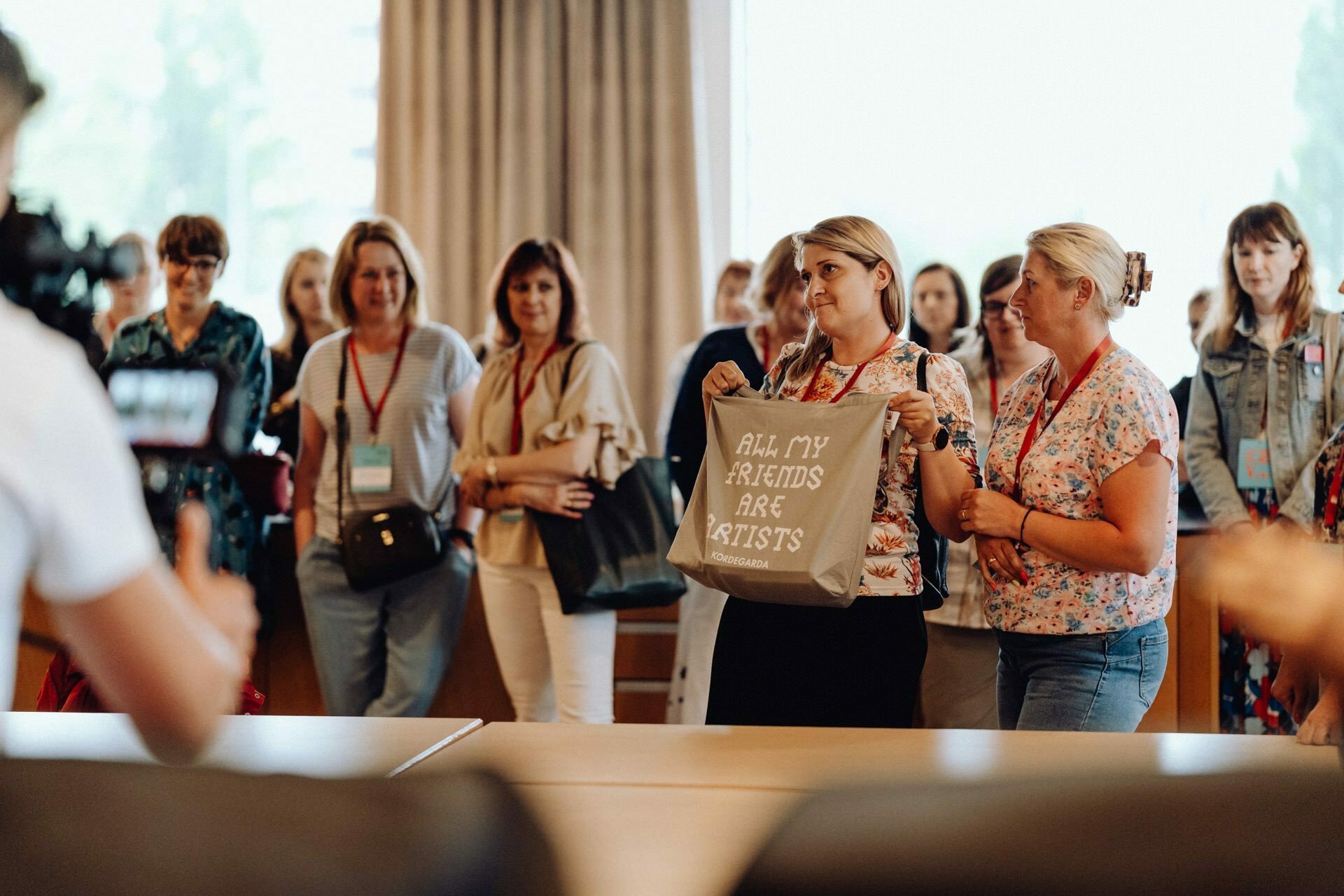 A group of people are standing attentively in a well-lit room, probably at a workshop or seminar. In the foreground, a woman holds a gray bag with the caption "ALL MY KNOWLEDGES ARE ARTISTS," while a photographer from Warsaw captures this moment of photojournalism from the event. 