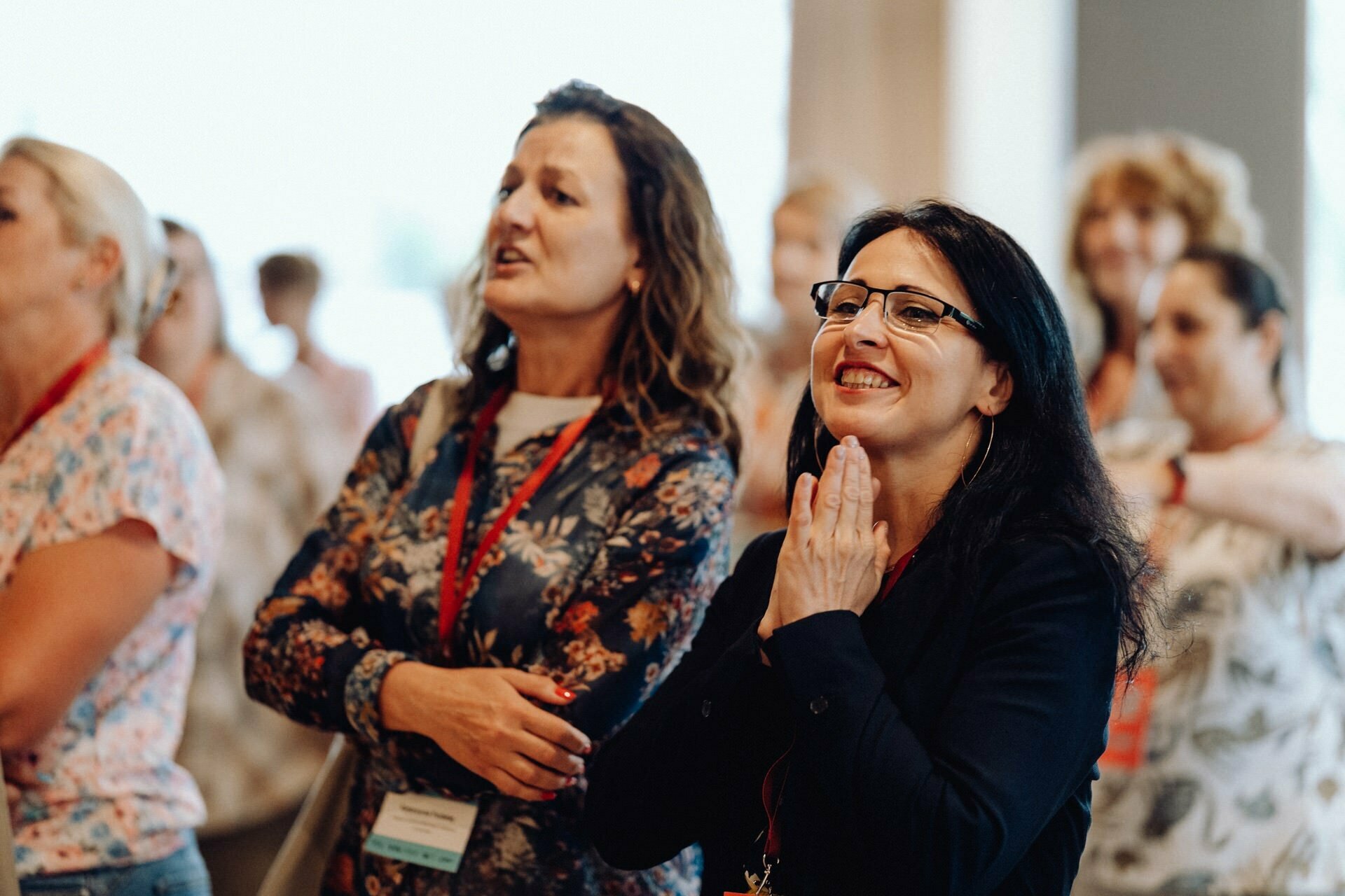 A group of women have gathered in the room, smiling and looking engaged. Some are wearing lanyards with name badges. The woman in the foreground, wearing glasses and dark hair, weaves her hands under her chin. The atmosphere seems lively and positive, which was perfectly captured by the photographer for the Warsaw event.   