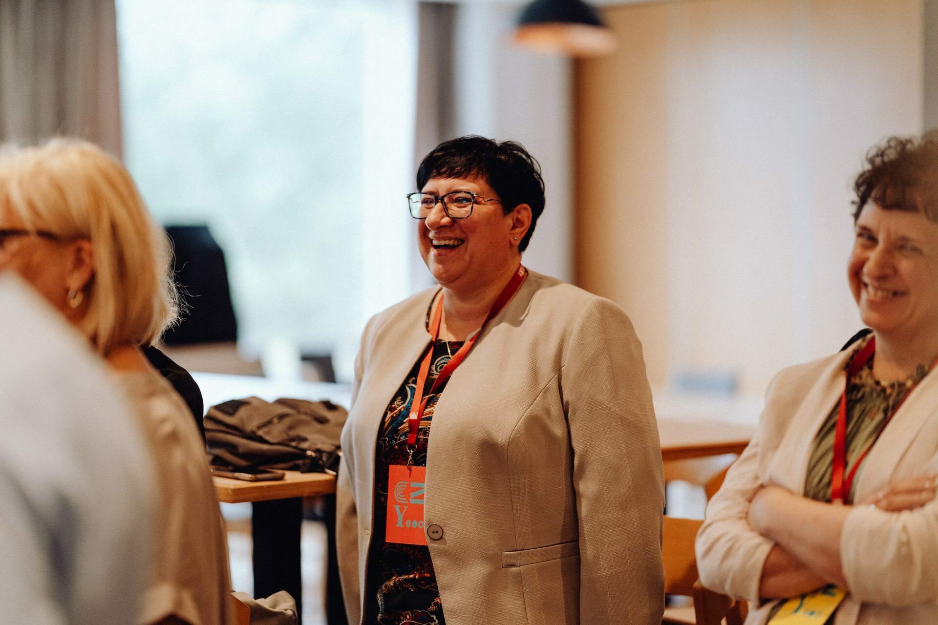 A group of smiling and laughing people in a well-lit room, perfectly captured by a photographer for an event. They are casually dressed, and the blurred background elements suggest they are in a social or professional gathering place. The room is equipped with natural light and wooden furniture.  