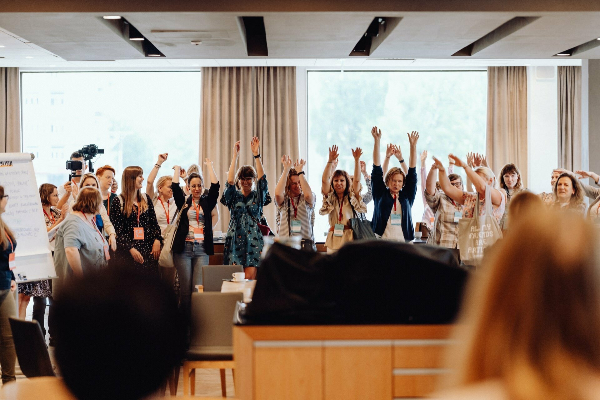 A group of people stand in a semicircle in a bright room with large windows. They raise their hands and smile, apparently engaged in a collective action. Several people are wearing badges, and to the left is a visible camera operator, perhaps a photographer at the event from Warsaw.  