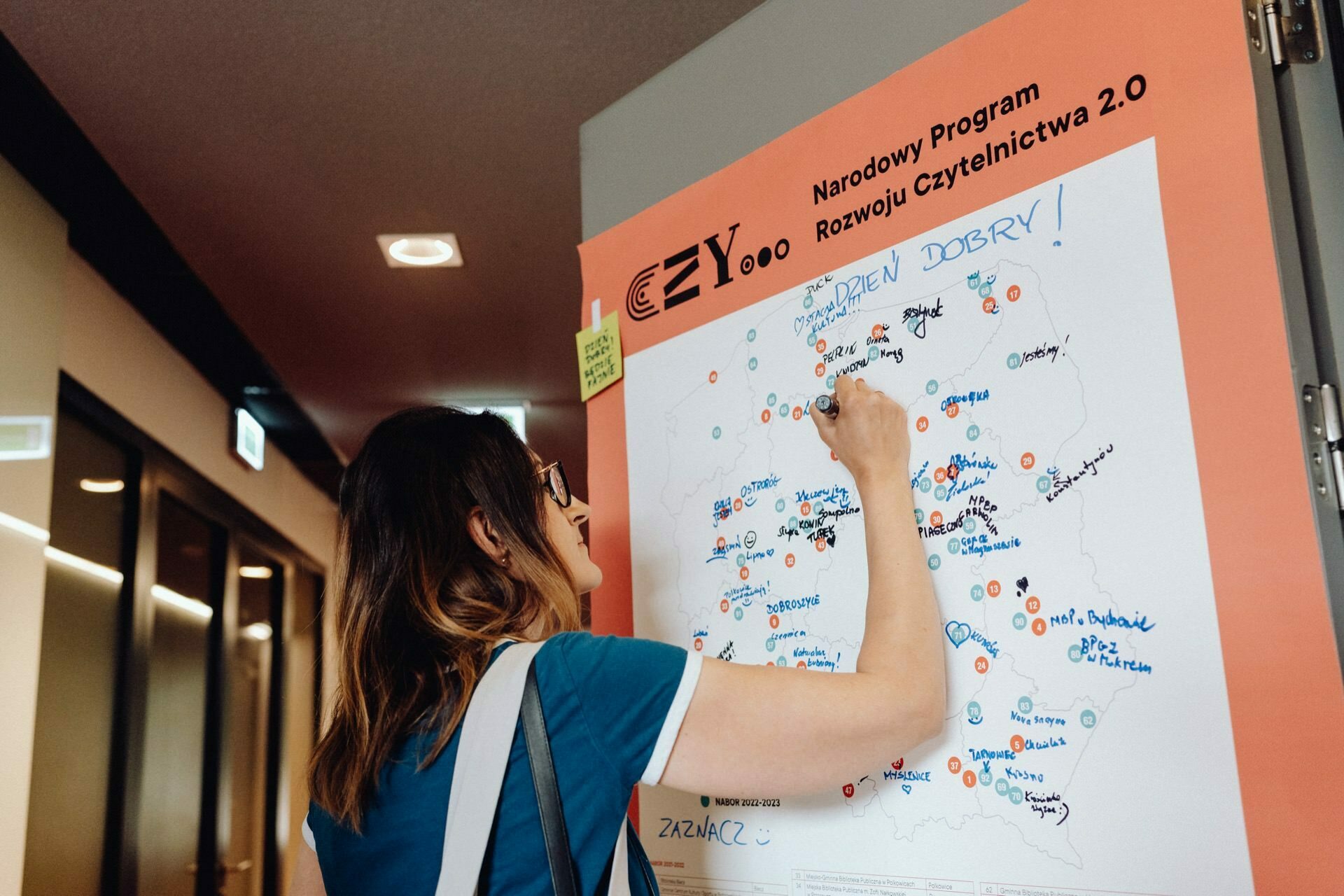 A woman with glasses and brown hair is writing on a large map posted on a board with the words "National Reading Development Program 2.0." The map features colorful markings and names indicating various locations, captured vividly by an event photographer warszawa, ensuring that every detail is preserved. 