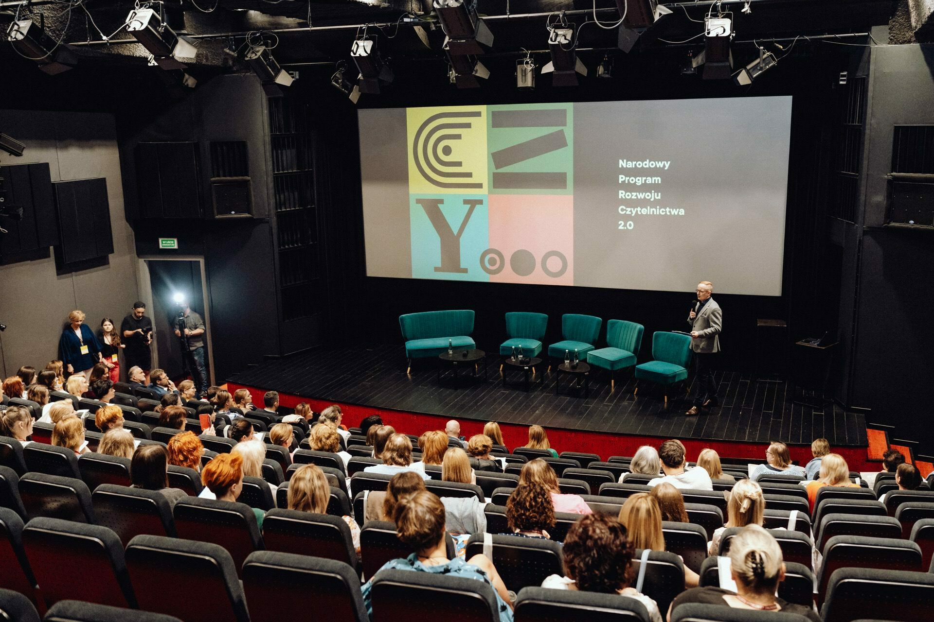 In a conference room filled with an audience seated facing the stage, five turquoise chairs are set up next to a podium on which the speaker stands. Behind them, "National Reading Development Program 2.0" is projected on a large screen, along with colorful abstract designs, perfect for photo coverage of the event by any event photographer. 