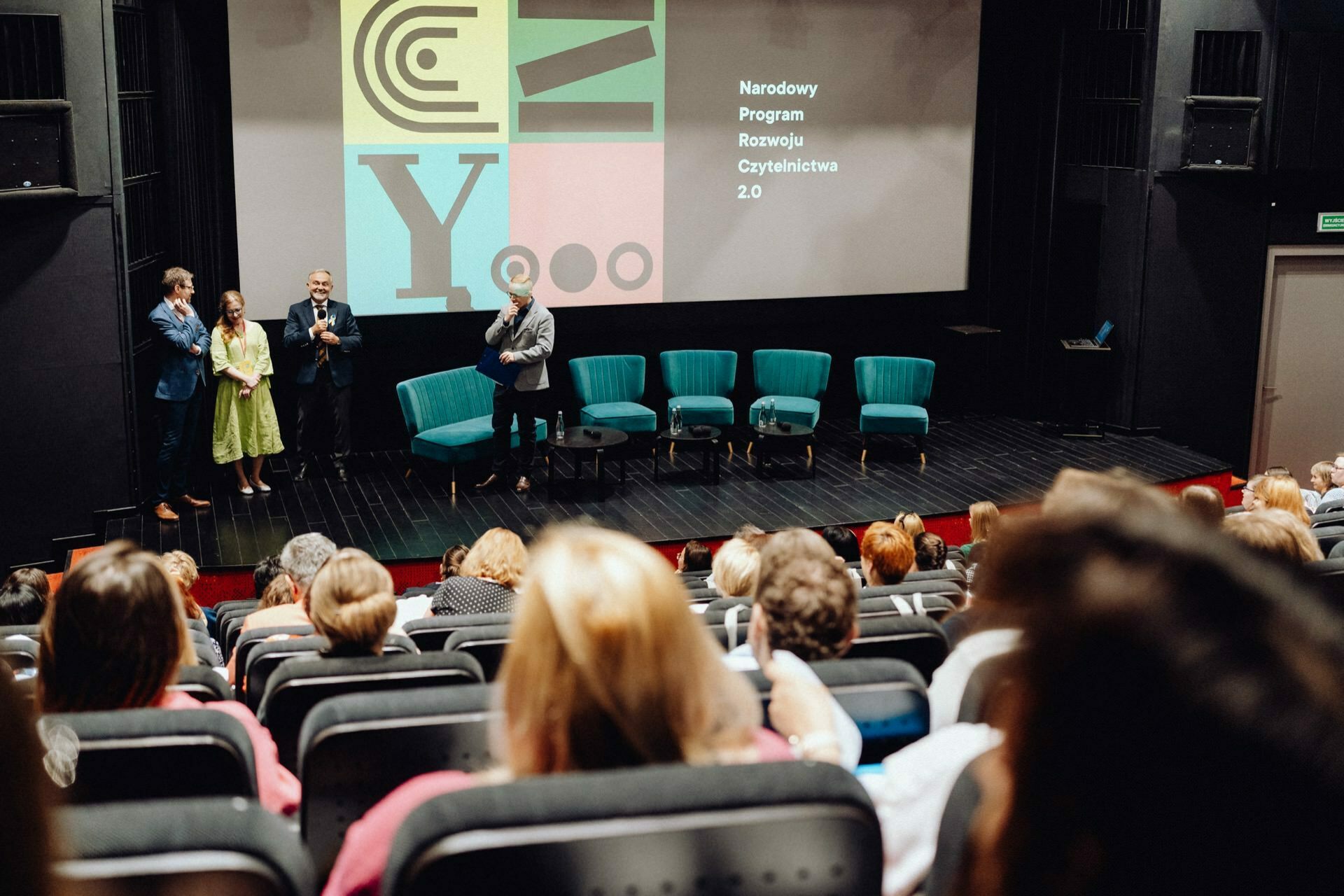 A group of people stand on a stage in front of a seated audience in a theater. The stage has a large screen displaying text and colorful graphics. Six empty turquoise chairs are placed on the stage. Attendees face the presenters, and a photographer at the event captures every moment, providing detailed photo coverage of the event.   