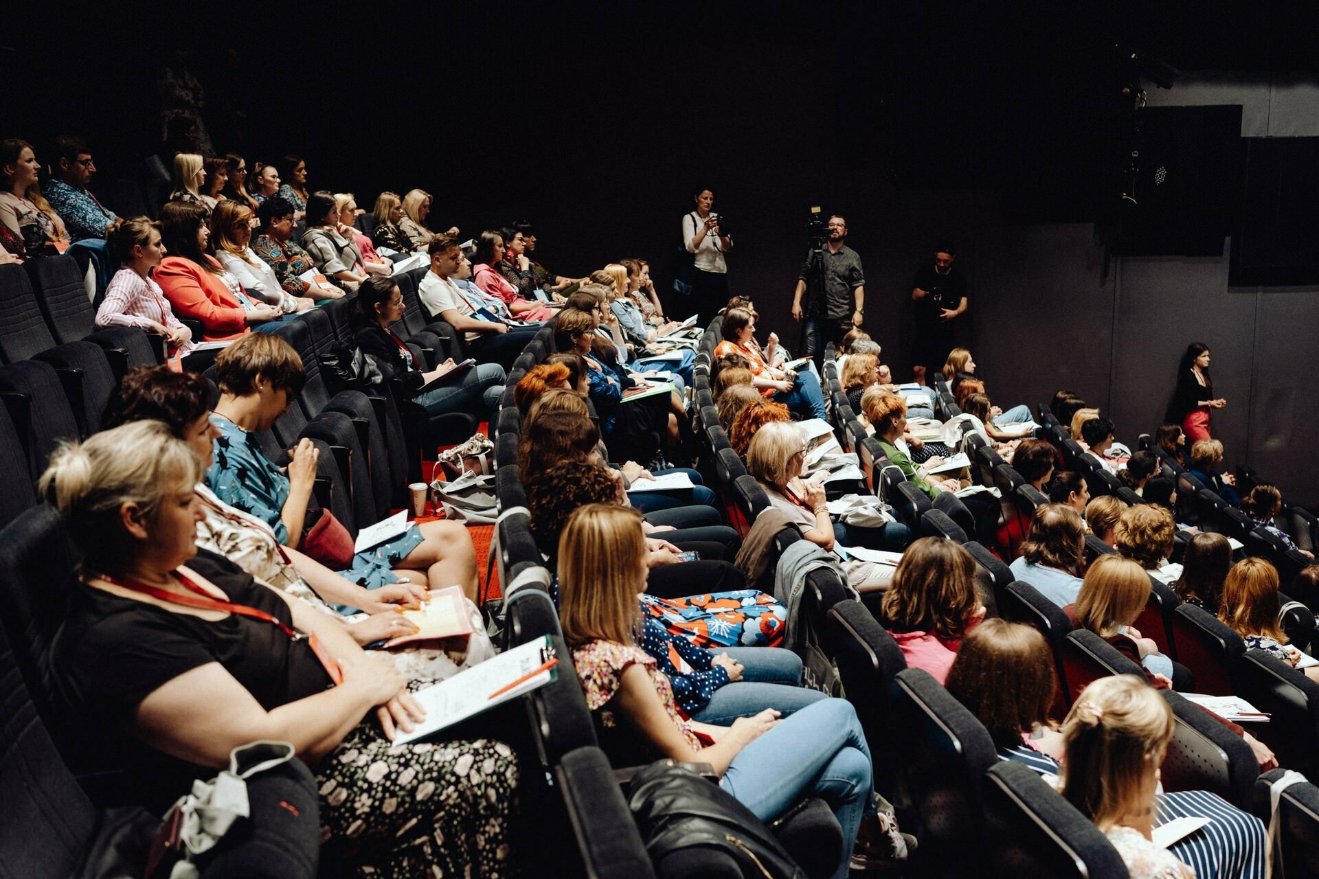 The large audience sits in tiered seats and carefully watches the speaker, who is out of frame. Many attendees are taking notes, and the crowd is diverse in age and appearance. The setting is reminiscent of a lecture hall or auditorium with dim lighting, ideal for detailed photo coverage of the event.  