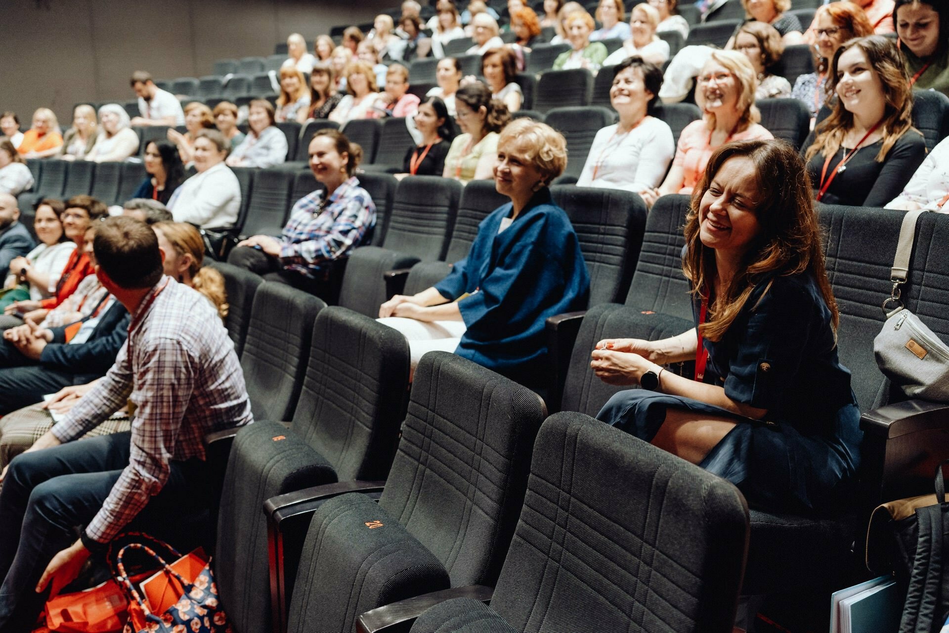 A large group of people sit in an auditorium with tiered seating, facing forward. Many people are smiling or laughing, creating a lively and engaging atmosphere. The audience consists of a variety of people of different ages and backgrounds, all of whom were beautifully captured by the photographer at the Warsaw event.  