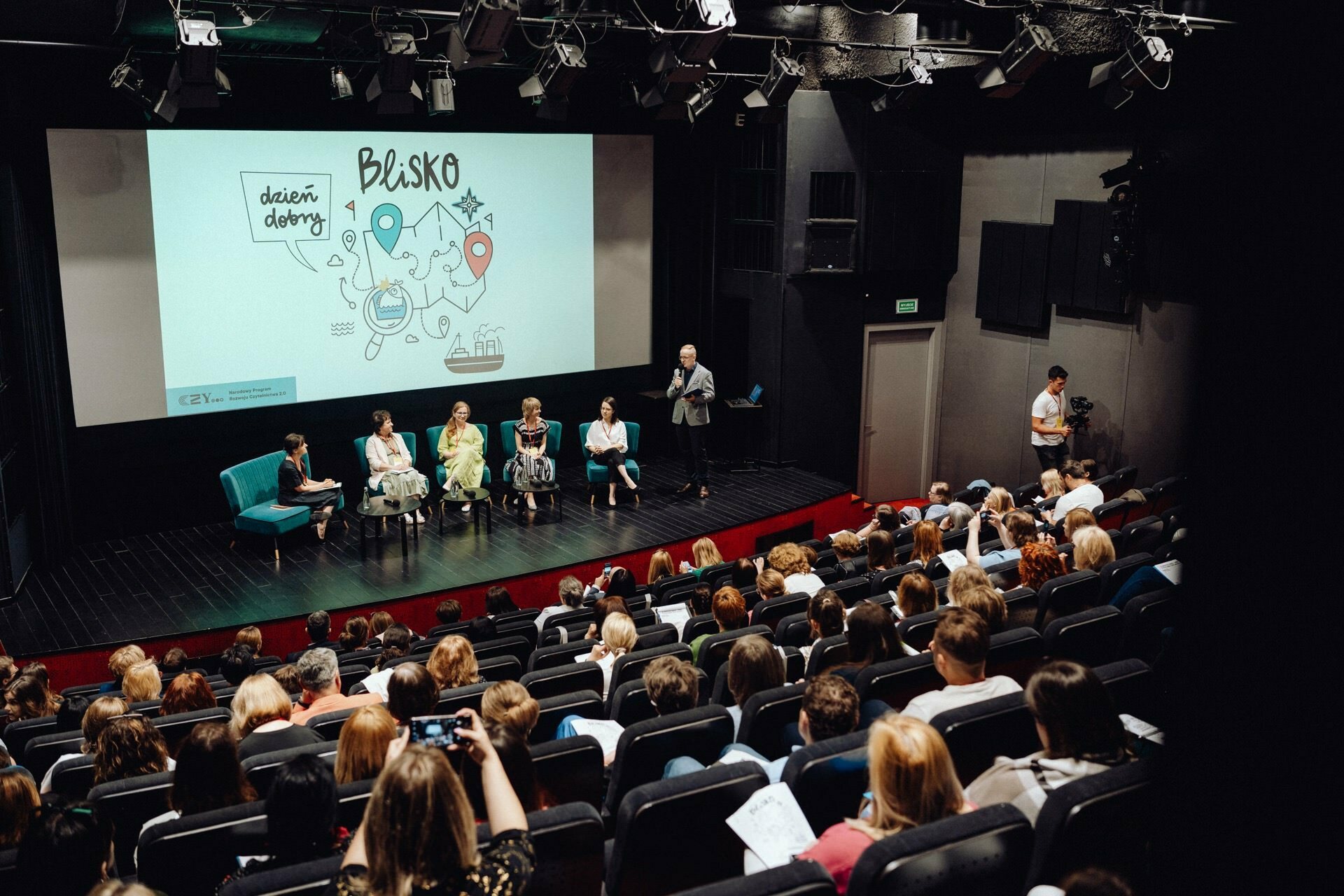 The audience is seated in the theater, facing the stage, where there is a large screen on which a drawing is projected, along with the words "good morning" and "BLISK". Six people sit on the stage, one stands at the lectern, while another, presumably a photographer from Warsaw, operates a camera near the exit. 