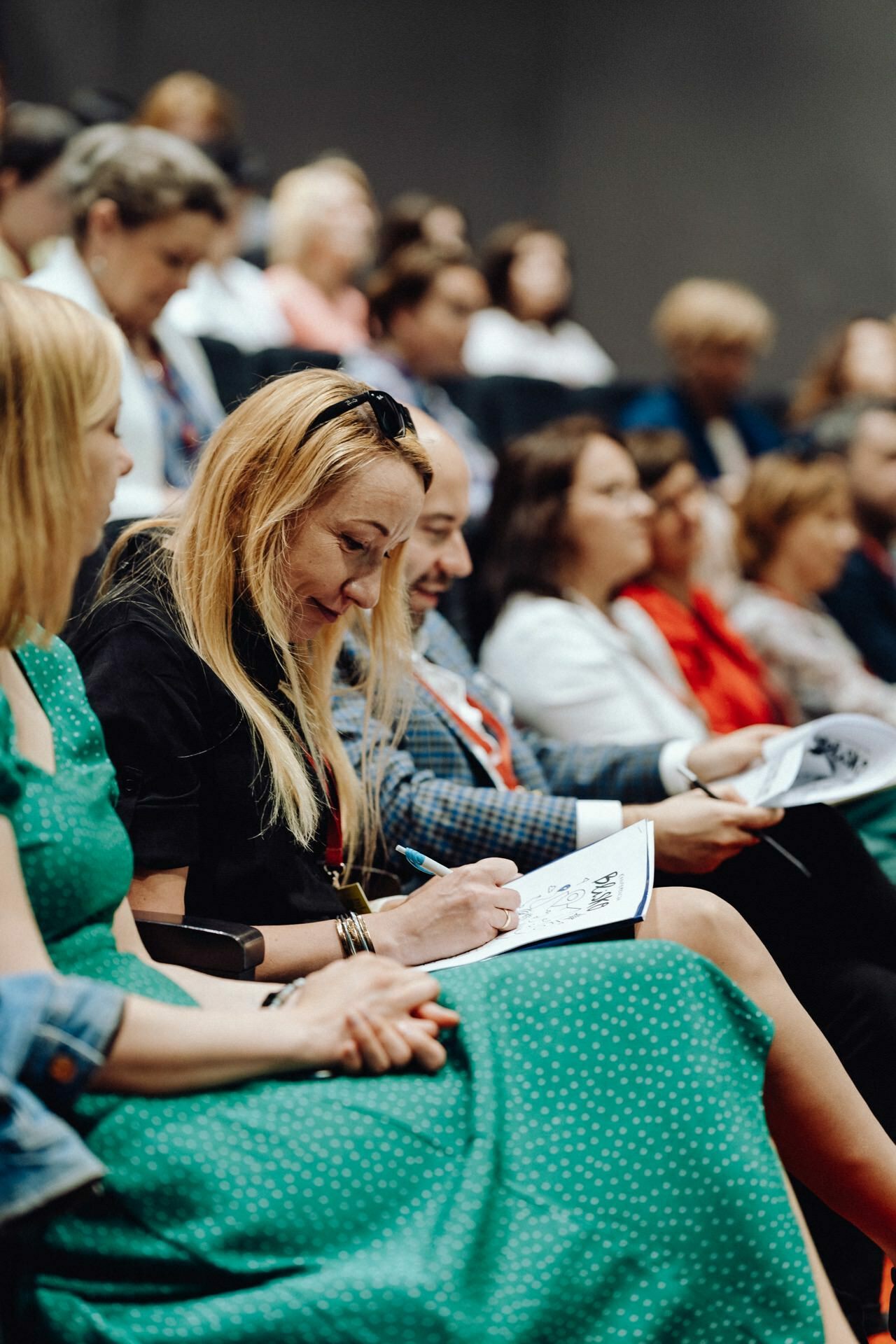 The woman in the foreground, wearing a green dress, sits among a group of people in an auditorium. She writes in a notebook and smiles. Others around her are sitting, some concentrating on their notes, others listening intently - an ideal scene for a photo essay on an event by any experienced photographer at an event. The background is slightly blurred.   