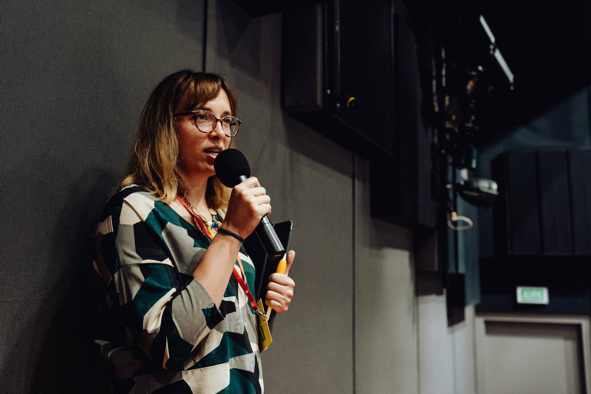 A person with glasses and shoulder-length hair, probably an event photographer Warsaw, is speaking into a microphone in a dimly lit room. They are wearing a patterned shirt and holding a book or notebook. Acoustic panels cover the walls, and audiovisual equipment is visible in the background.  