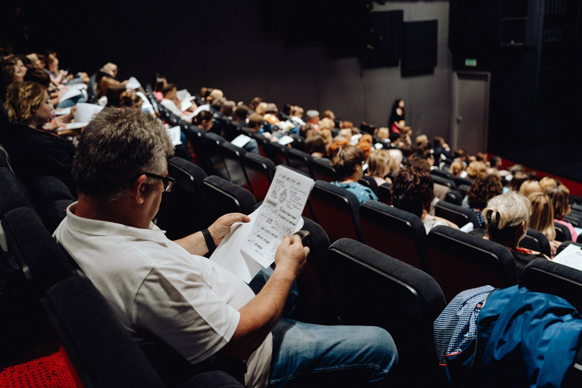 A man in a white shirt sits in a crowded room and reads a piece of paper covered with handwritten notes. The floor seats are filled with other people busy with various activities, and at the back of the room stands a person capturing moments for a photo essay of the event by a photographer from Warsaw. 