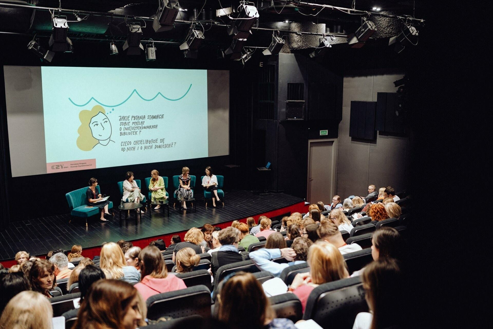 A conference room with a seated audience facing the stage where four panelists are discussing. Behind them, a large screen displays an illustration and text. The room is dimly lit and the panelists are seated in blue chairs. A photographer from Warsaw meticulously captures the event for a photo report of the event.   