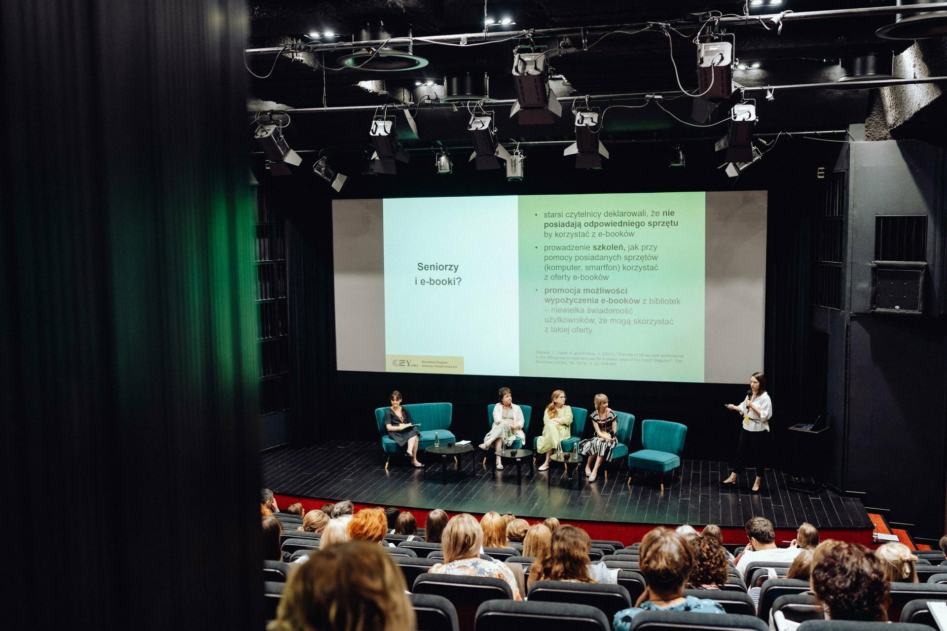 A group of four people sit on a stage in an auditorium, facing the audience. They are taking part in a presentation with a screen displaying a slide entitled "Seniors and e-books?" A person, presumably the presenter, stands near the screen while the audience listens intently. This scene is ideal for an event photographer Warsaw to capture moments for an event photo report.